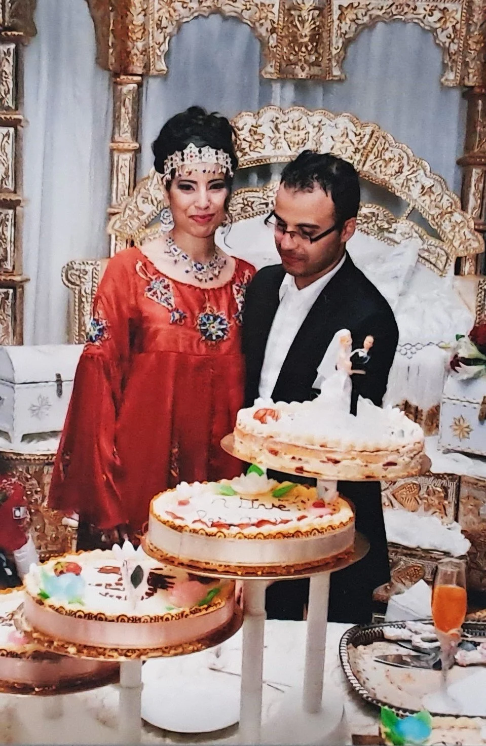 Un couple lors d'une célébration de mariage avec des gâteaux décorés sur une table. La femme porte une robe rouge avec des broderies et un diadème, l'homme porte un costume noir avec une chemise blanche. La scène est décorée avec des éléments traditi