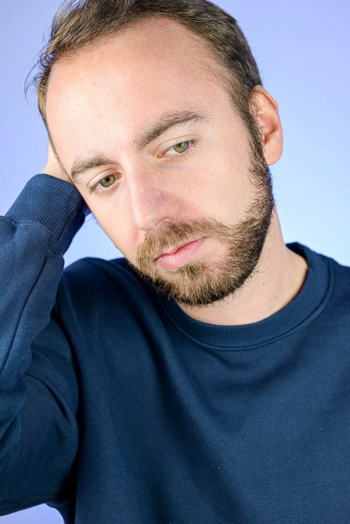 Acteur avec des cheveux et une barbe brune portant un sweat bleu clair, la tête penchée et l'air pensif, avec un fond bleu clair.