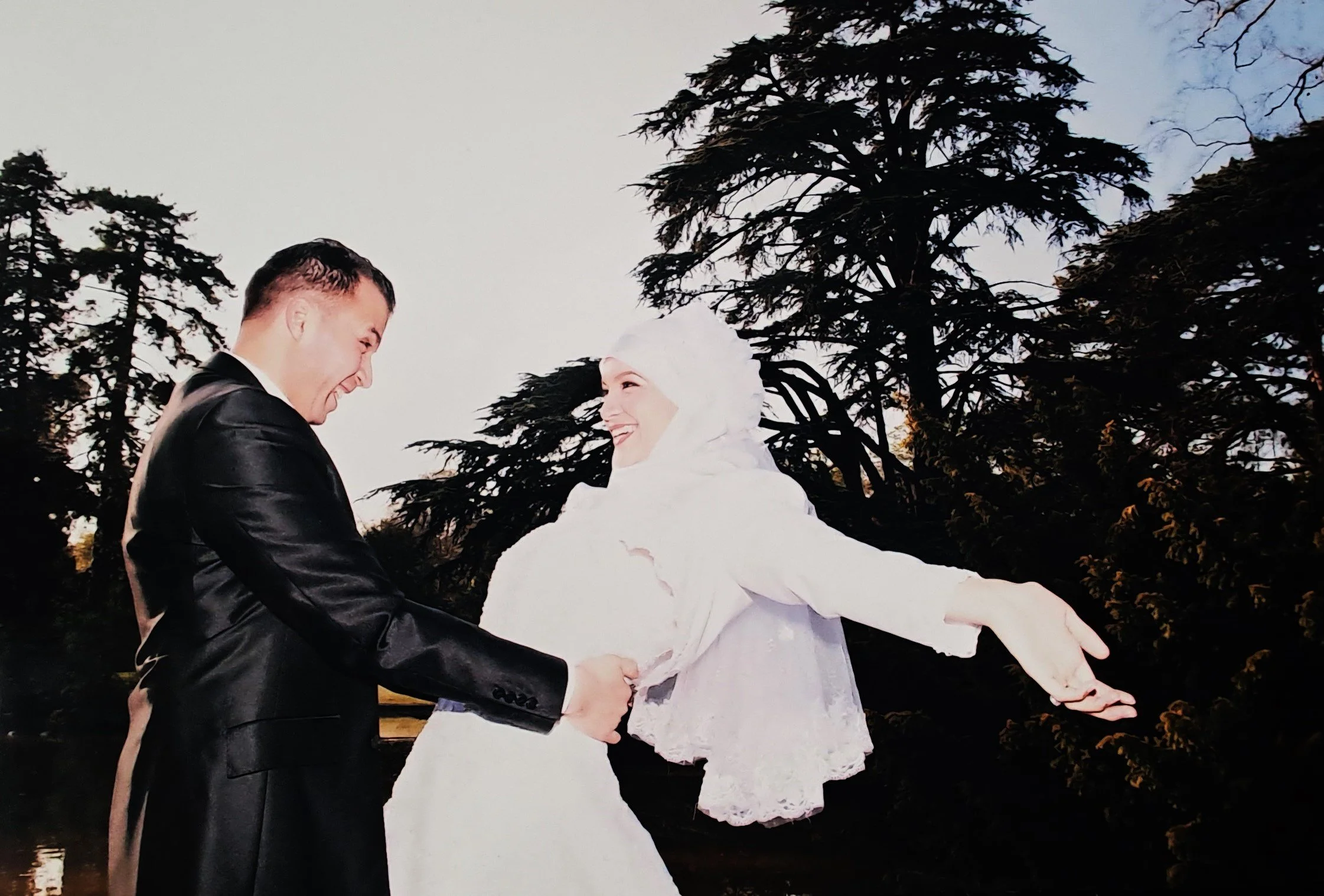 Un couple danse extérieure au coucher du soleil, avec des arbres en arrière-plan, la femme porte une robe blanche et un foulard, l'homme porte un costume noir.