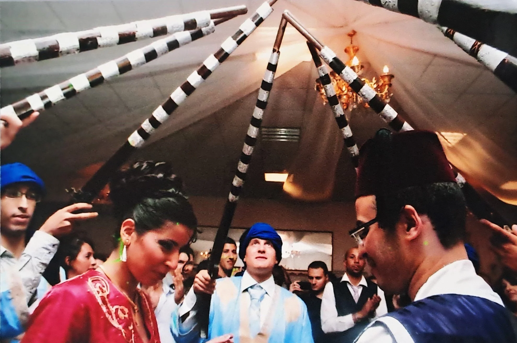 Groupe de personnes lors d'une célébration, certaines portant des chapeaux bleus, d'autres en tenue vestimentaire colorée, sous une tente décorée avec des chandeliers et des éléments décoratifs noirs et blancs.