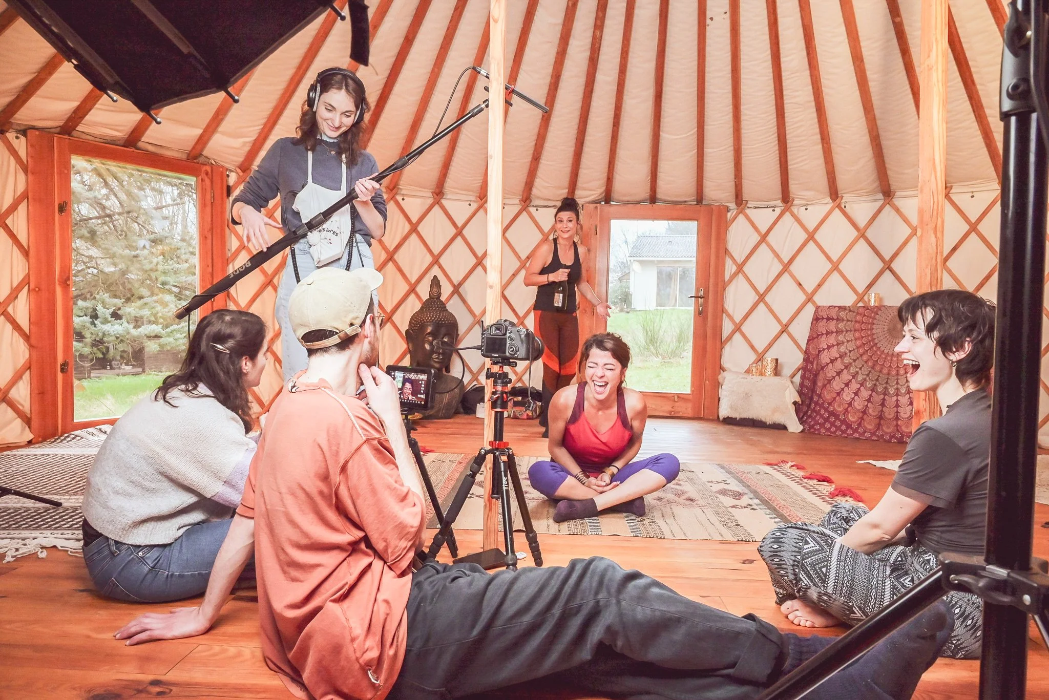 Groupe de personnes en train de tourner une vidéo ou un film dans une cabane en bois, avec une femme assise par terre riant et deux autres femmes en fond qui semblent expliquer quelque chose, caméra, micro et matériel de tournage présents.