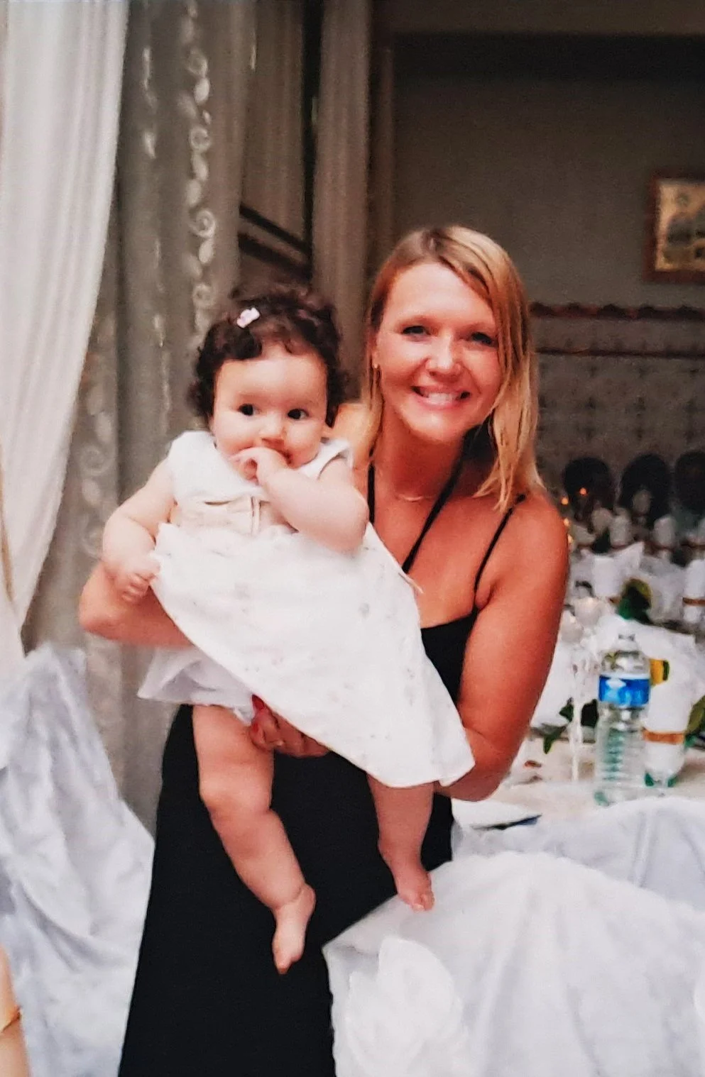 Une femme souriante tenant un bébé dans une pièce décorée pour un événement, avec une table dressée en arrière-plan.