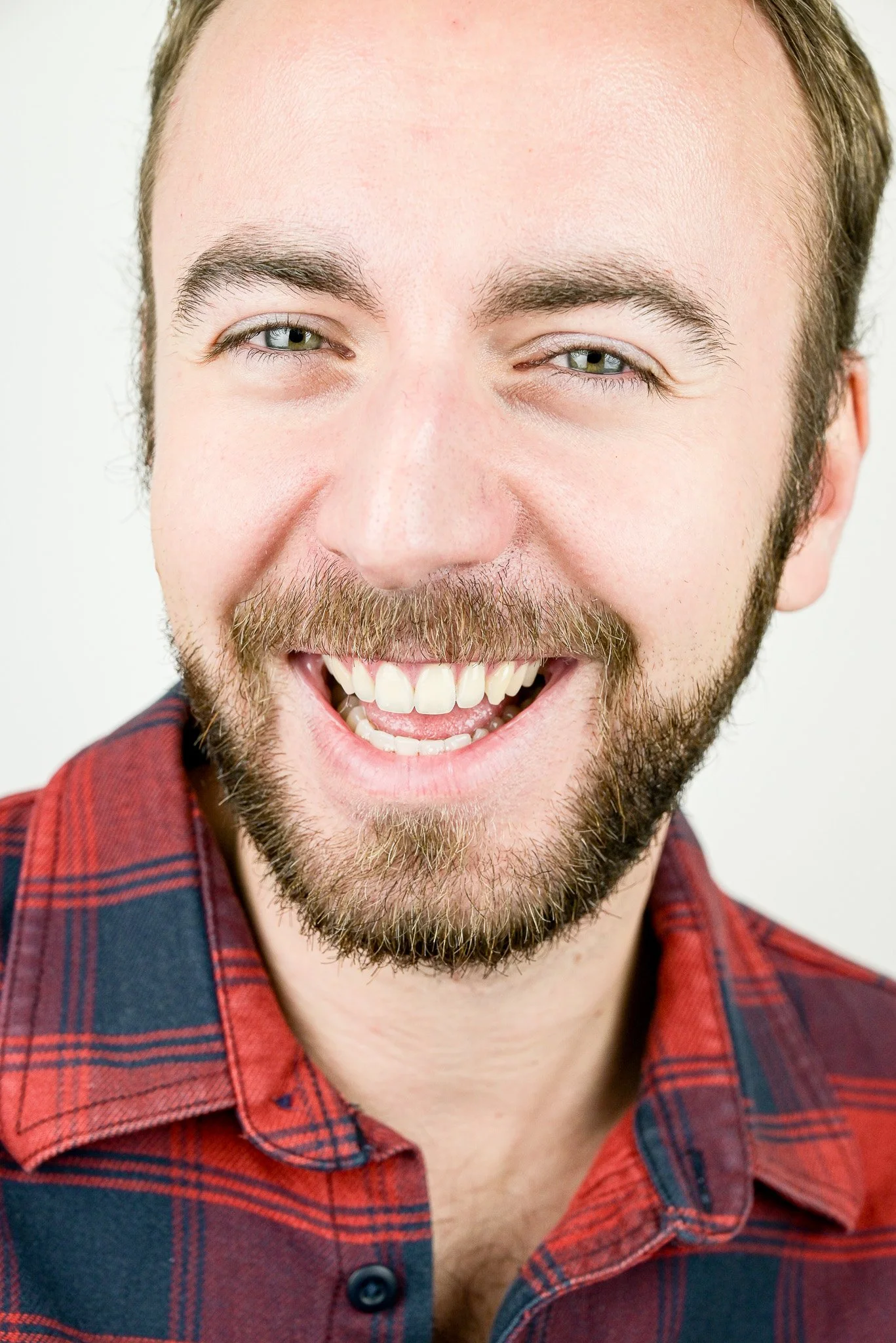 Acteur Thomas Kindler souriant avec une barbe, portant une chemise à carreaux rouges, aperçu en gros plan.