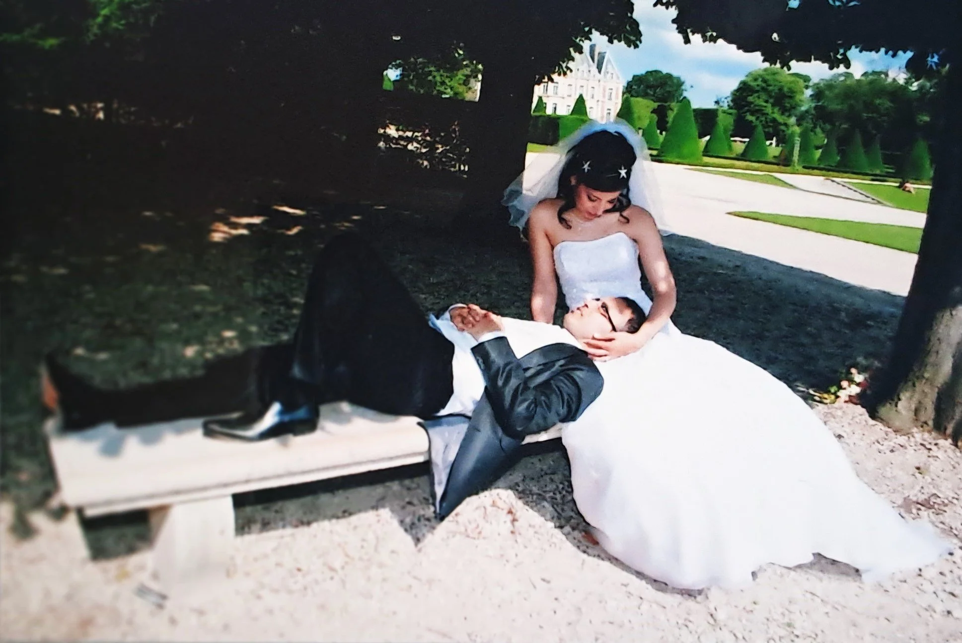 Une jeune femme en robe de mariage et un homme en costume allongés sur un banc dans un parc, elle le regarde tendrement.
