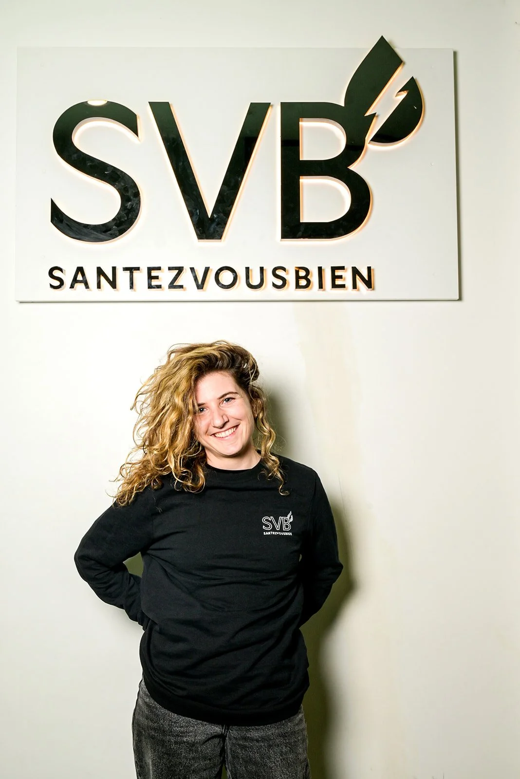 Séance photo portrait corporate. Jeune femme souriante en pull noir avec logo SVB Santezvousbien, debout devant un mur blanc avec un grand logo SVB et la mention SANTEZVOUSBIEN.