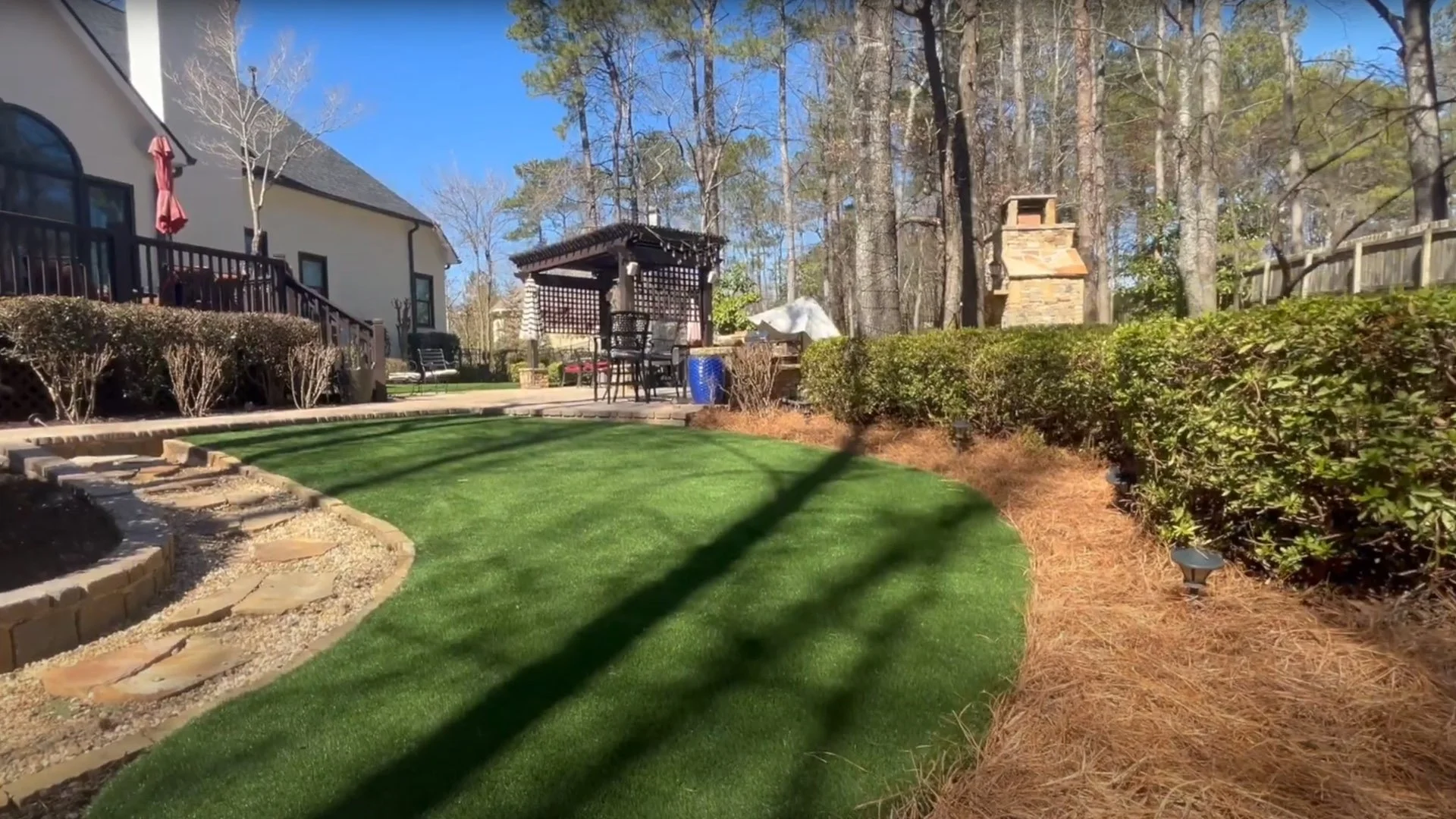 Backyard with green lawn, paved walkway, and pergola near a house, surrounded by trees and bushes.