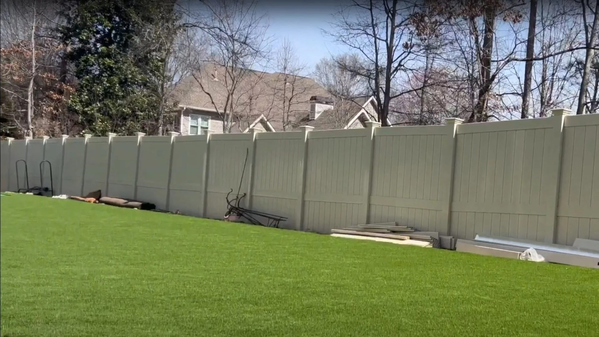 Backyard with green grass, white privacy fence, and scattered lawn equipment and materials.