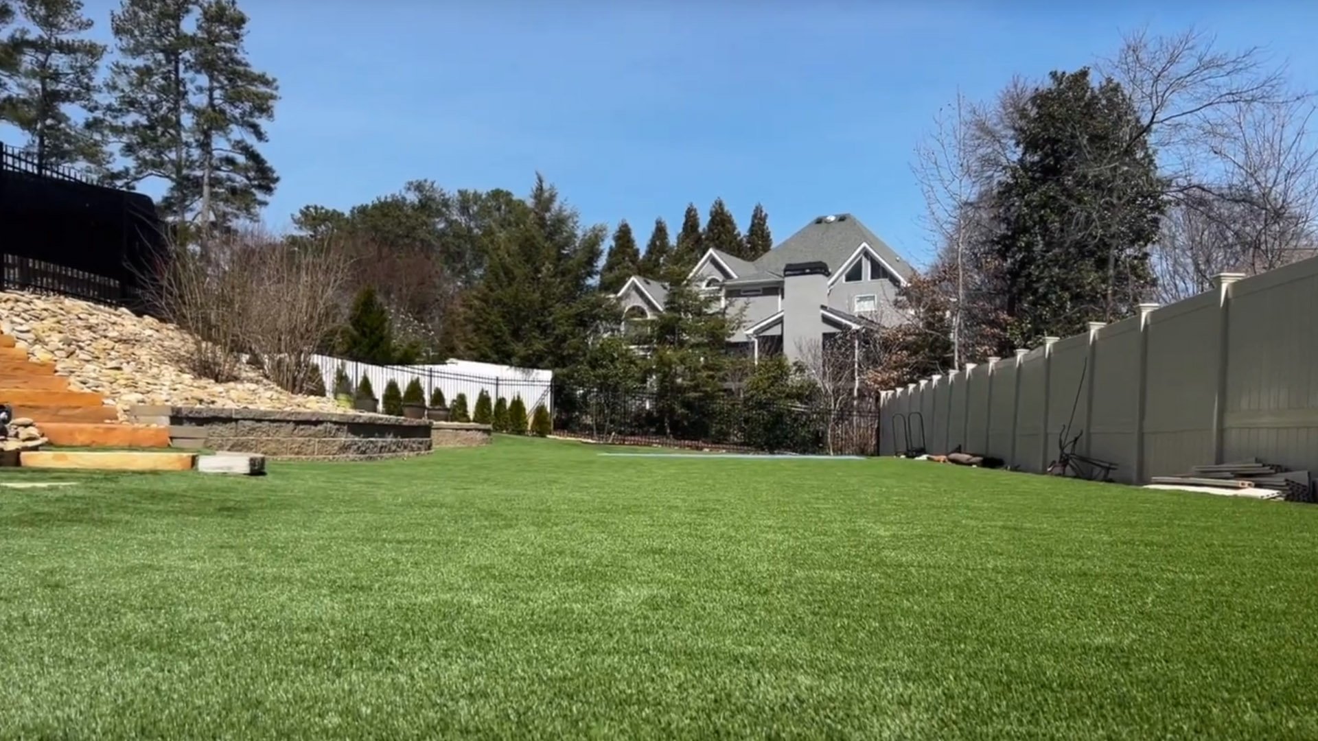 A lush green lawn with a modern house in the background, surrounded by trees and a white fence.