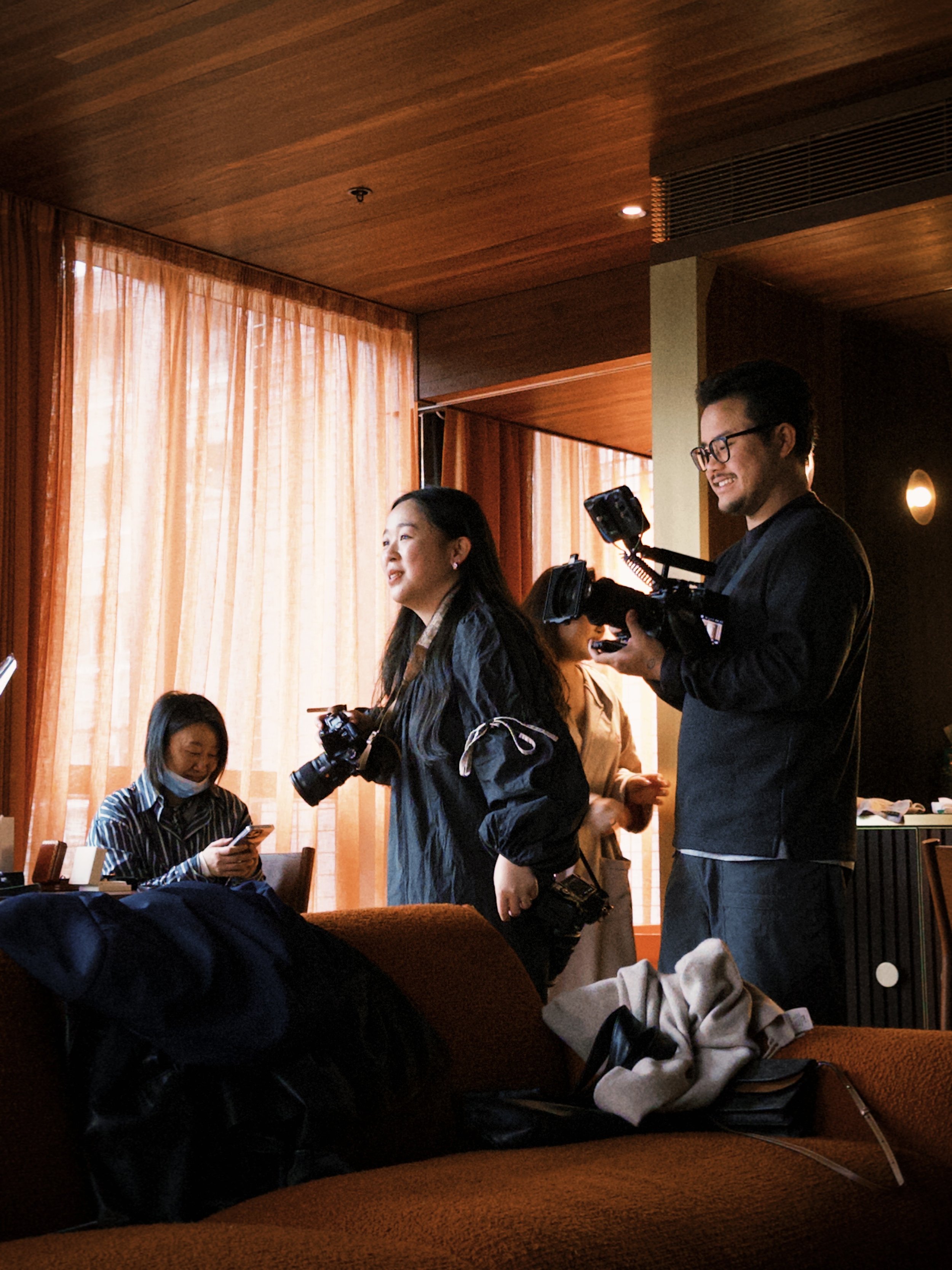 People gathered in a warmly lit room with wooden walls and large windows with curtains. One woman wearing a mask sits on a sofa using her phone, while a man with glasses showcases a camera and other women stand nearby, engaged in conversation.