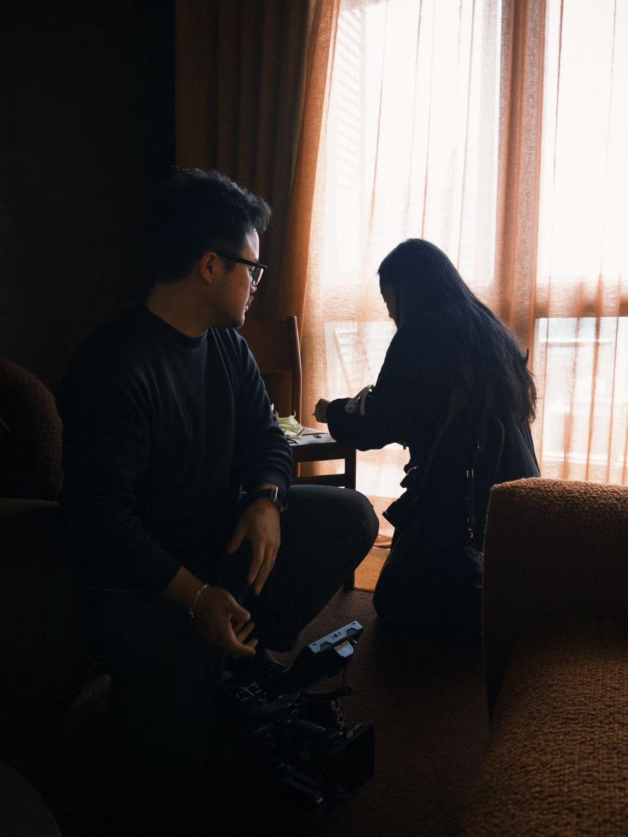 Two young people, a man and a woman, sitting in a dimly lit room near a window with curtains. The man is wearing glasses and a dark shirt, with a side profile visible. The woman has long dark hair, is looking down at her phone, and is dressed in dark clothing. There is a small table with items between them and a chair partially visible.