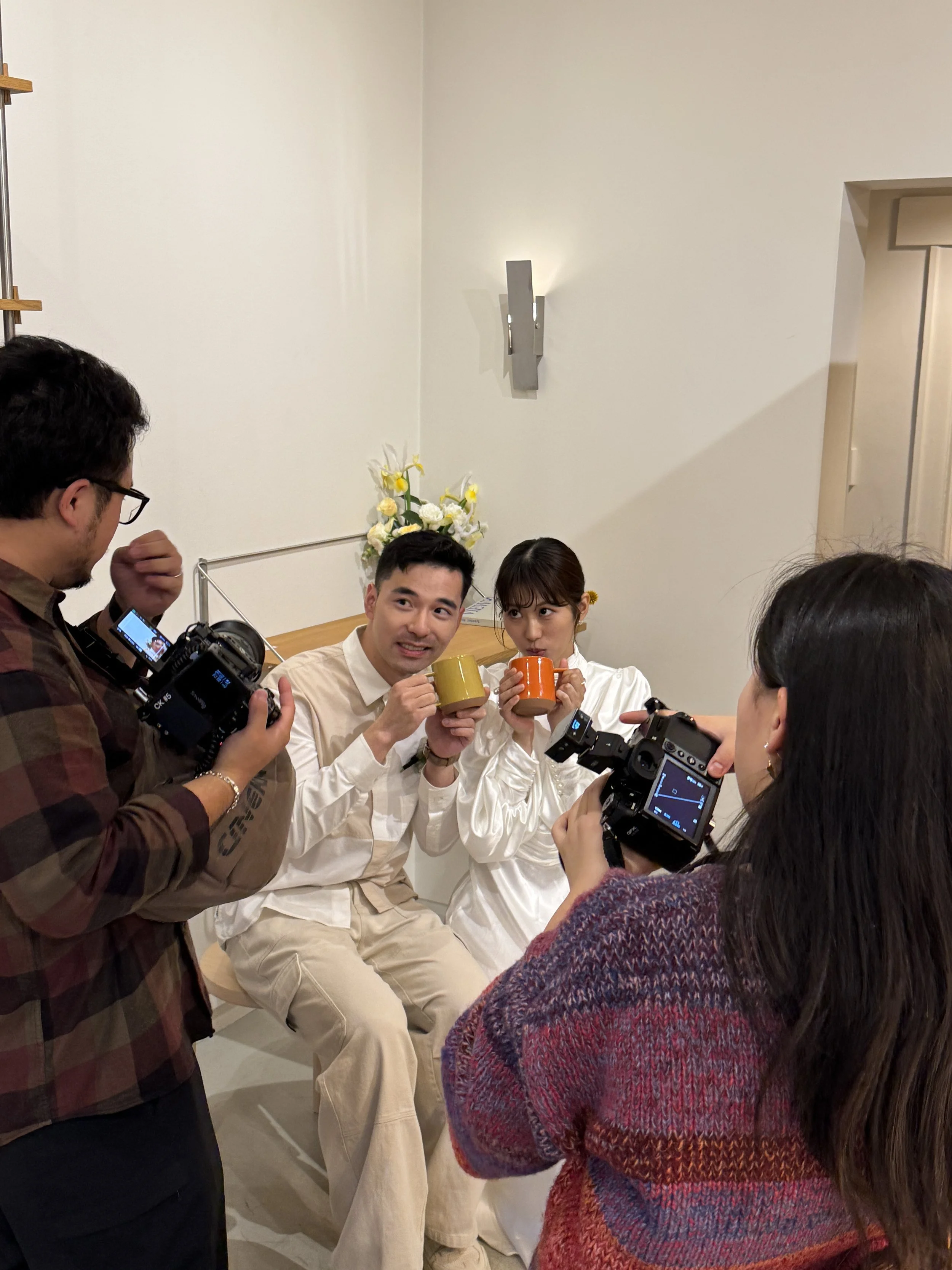 Group of people gathered around a man and woman holding coffee mugs, taking photos and selfies in a room with white walls and a floral arrangement in the background.