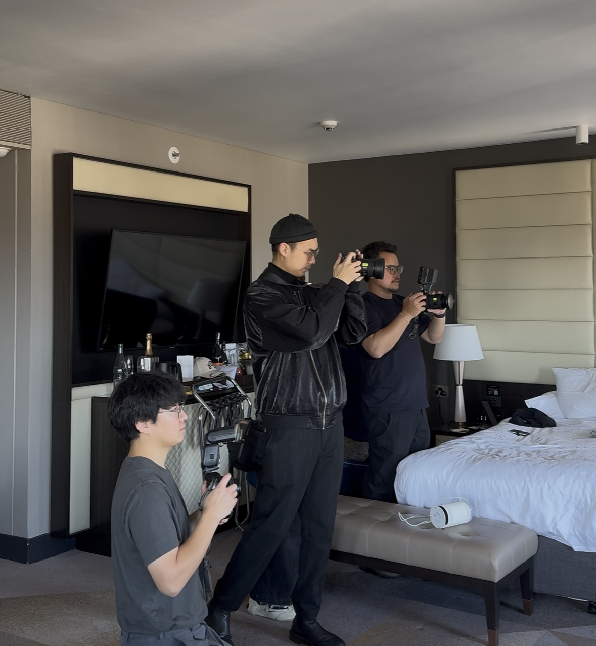 Three men with cameras filming inside a hotel room, with a bed, television, and other hotel furnishings visible.