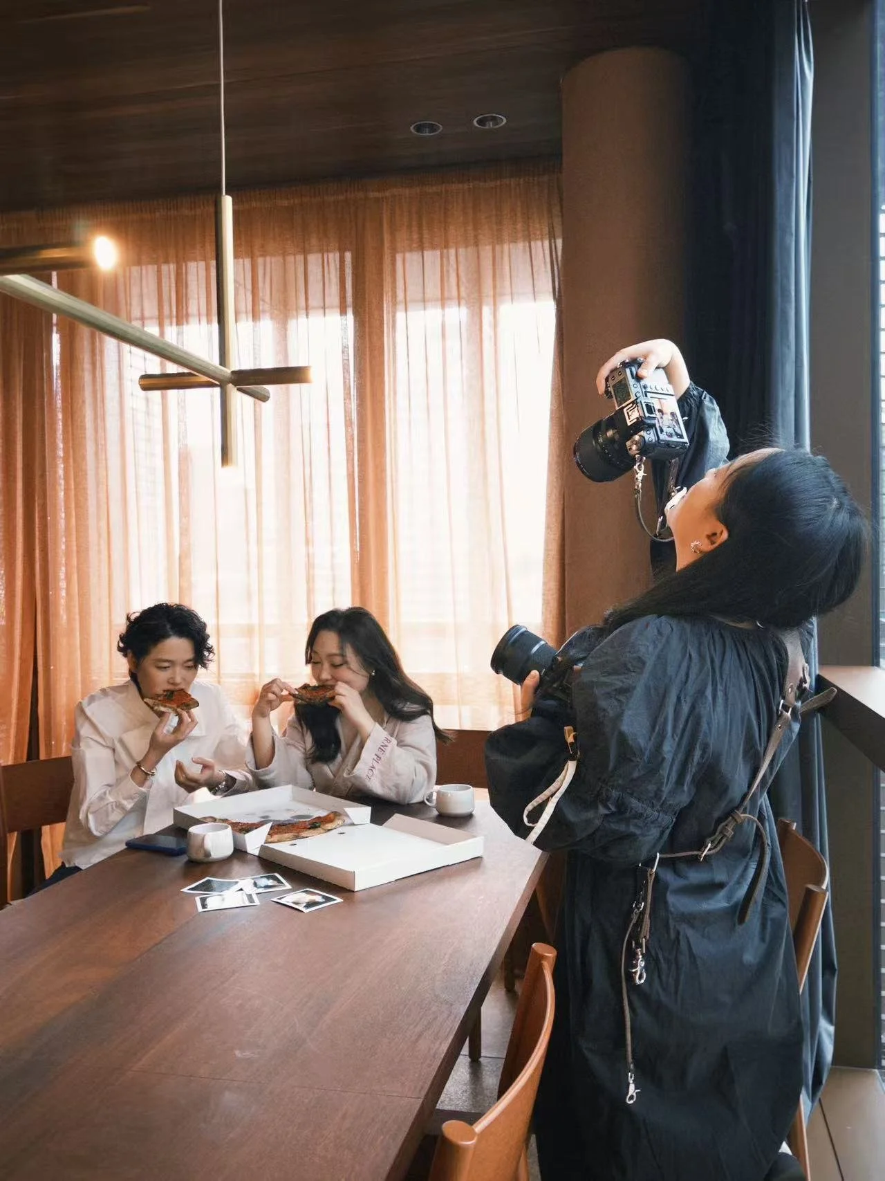 Two women sitting at a table in a restaurant, eating pizza, while a photographer takes photos of them.