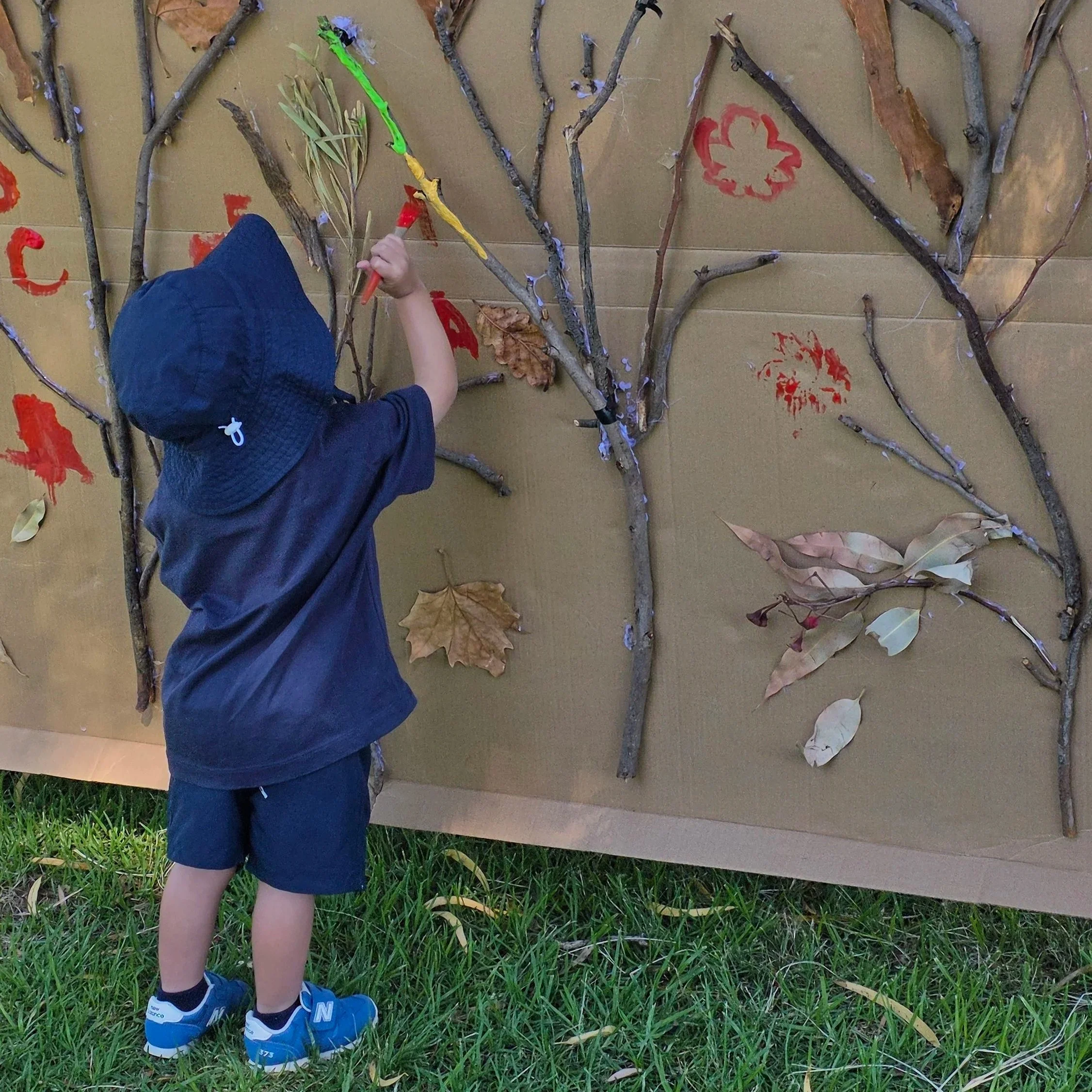 toddler painting outside, sticks and leaves stuck to cardboard at a messy play session