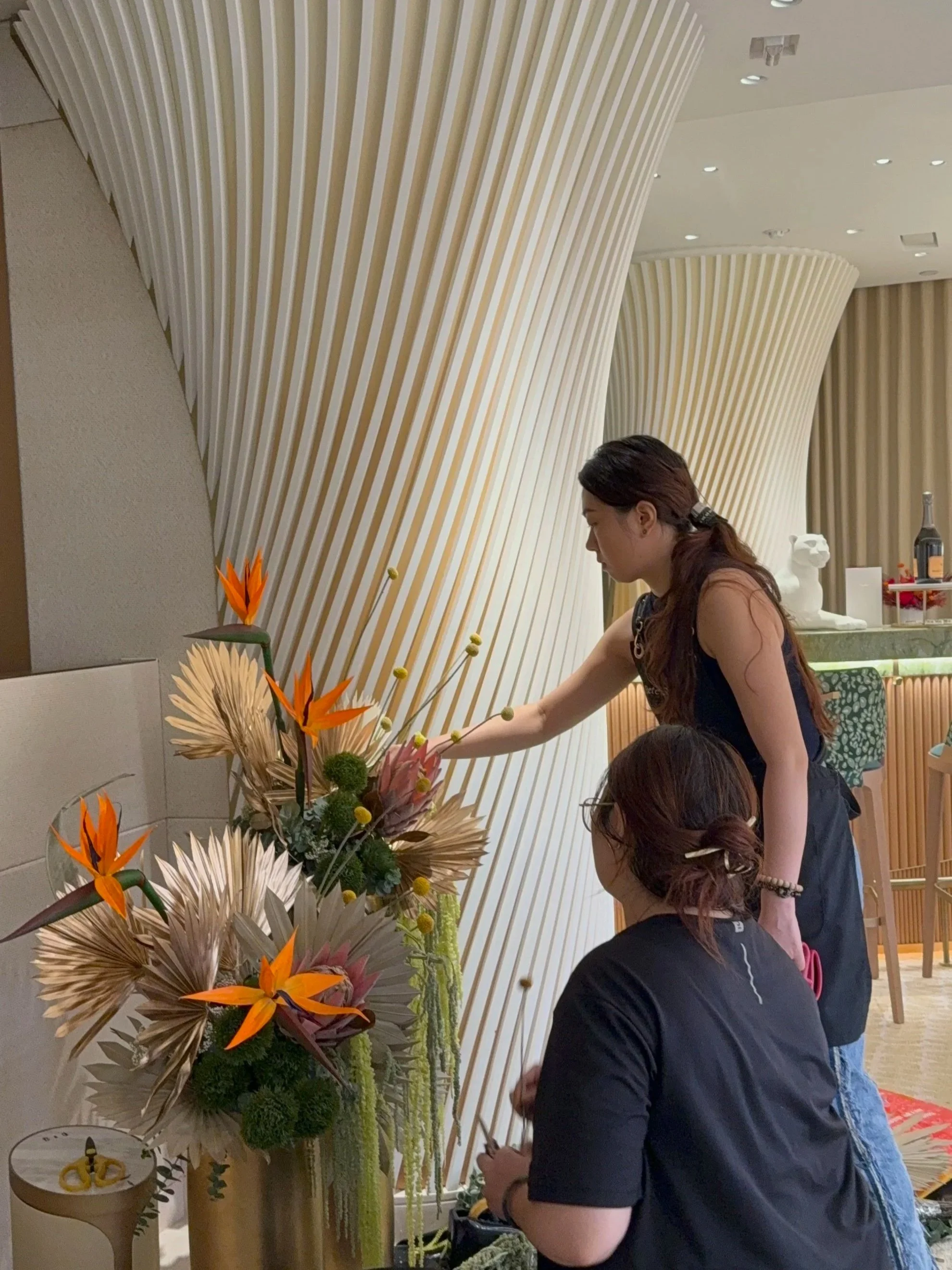Two women arranging a vibrant floral display with orange and beige flowers, set against a modern interior with curved, striped architectural elements.