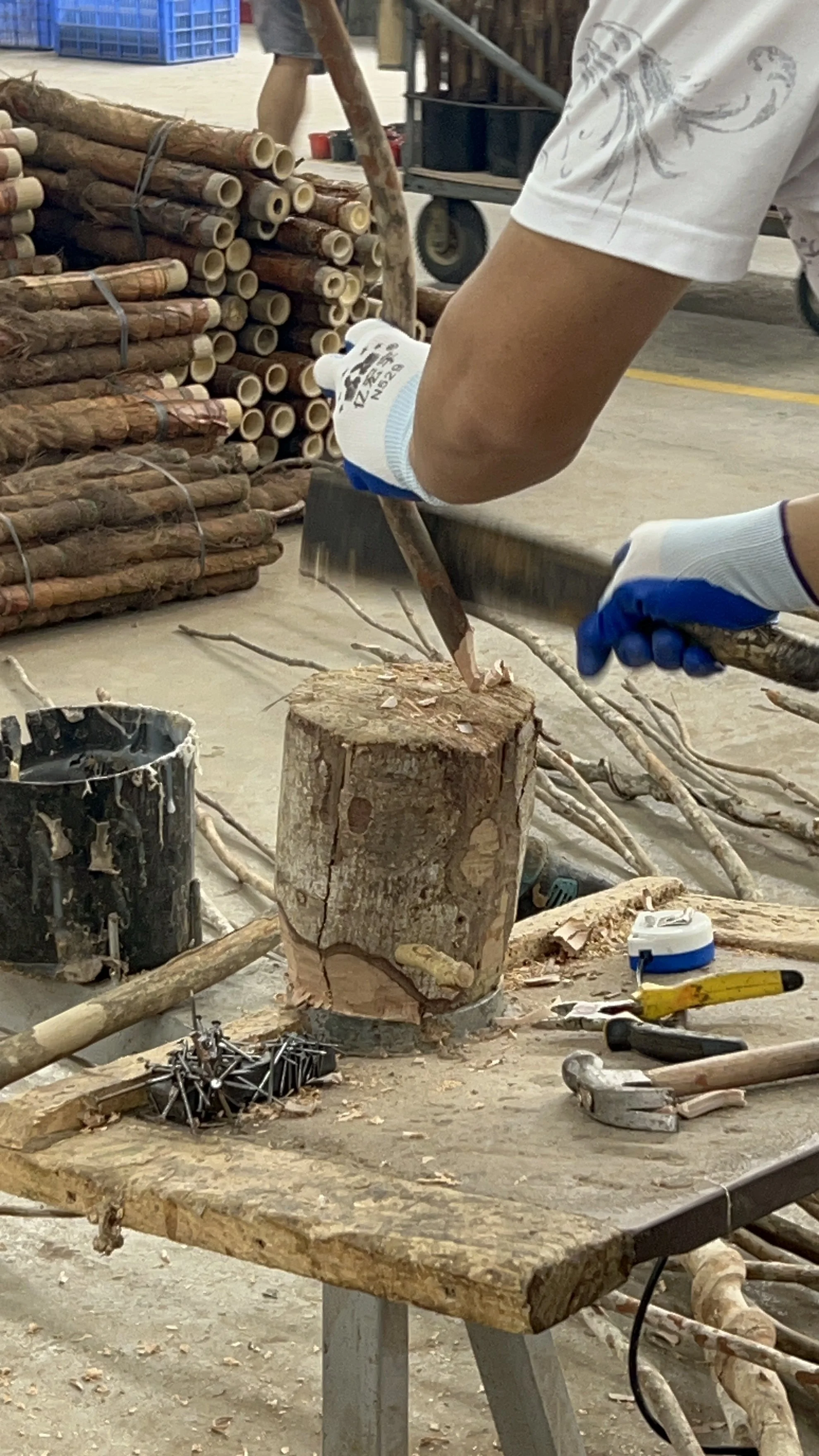 Person chopping wood with a hacksaw, surrounded by wood logs, tools, and wood shavings in a workshop or outdoor setting.