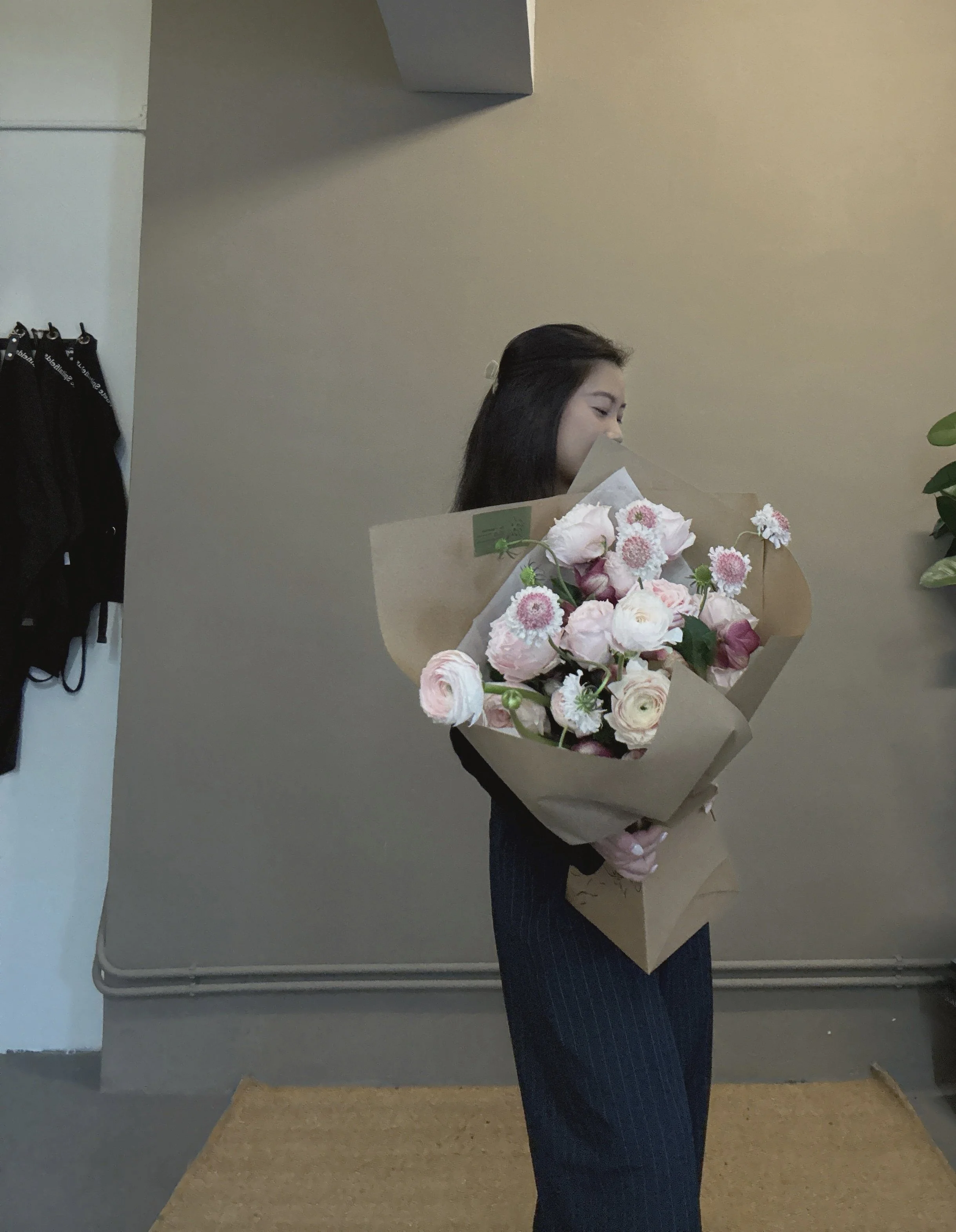 A woman holding a large bouquet of pink and white flowers, standing indoors with a neutral-colored wall behind her.
