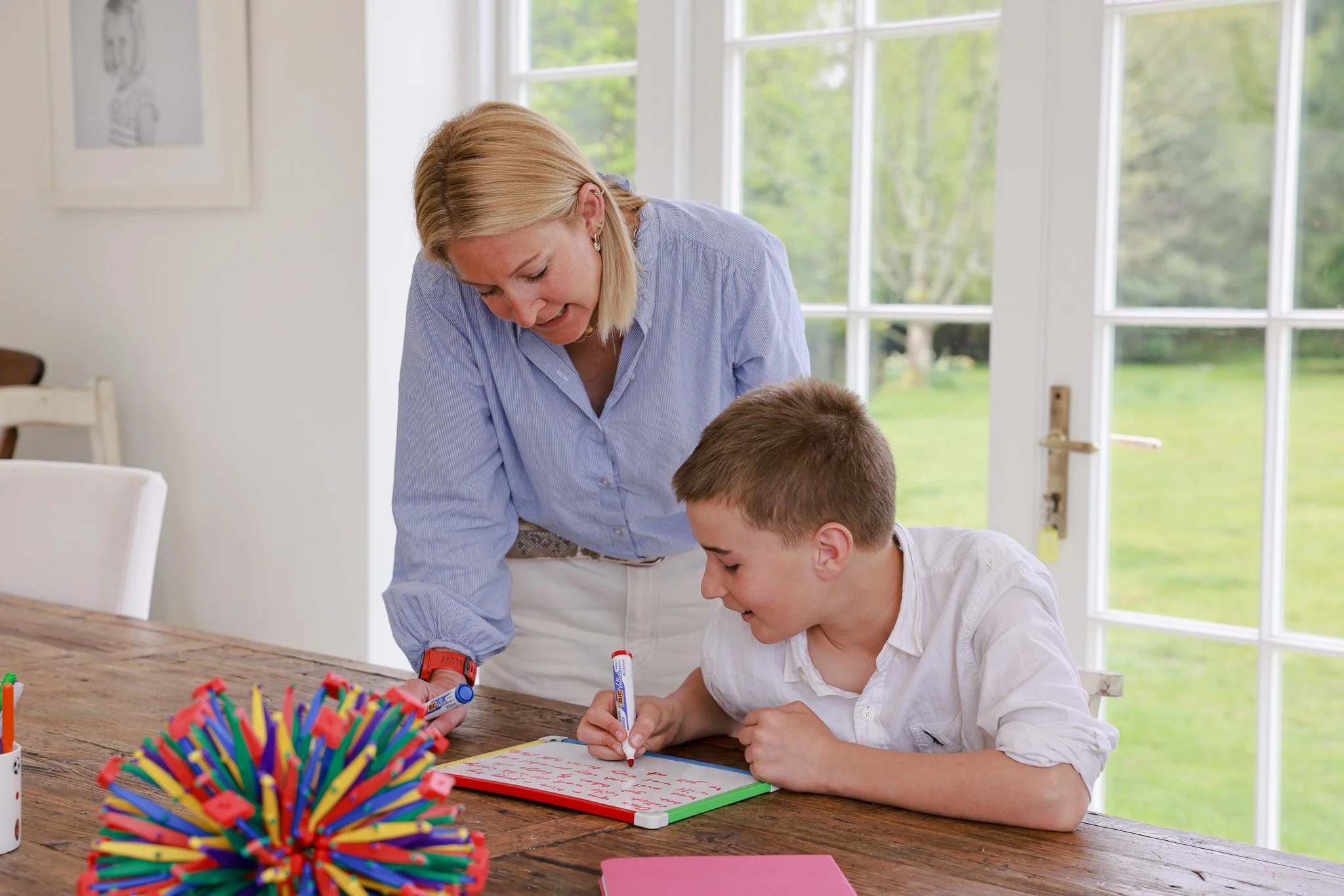 A woman and a boy sitting at a wooden table, writing on a whiteboard with dry erase markers, with colorful markers and a marker holder in the foreground, and a large window showing green trees outside.