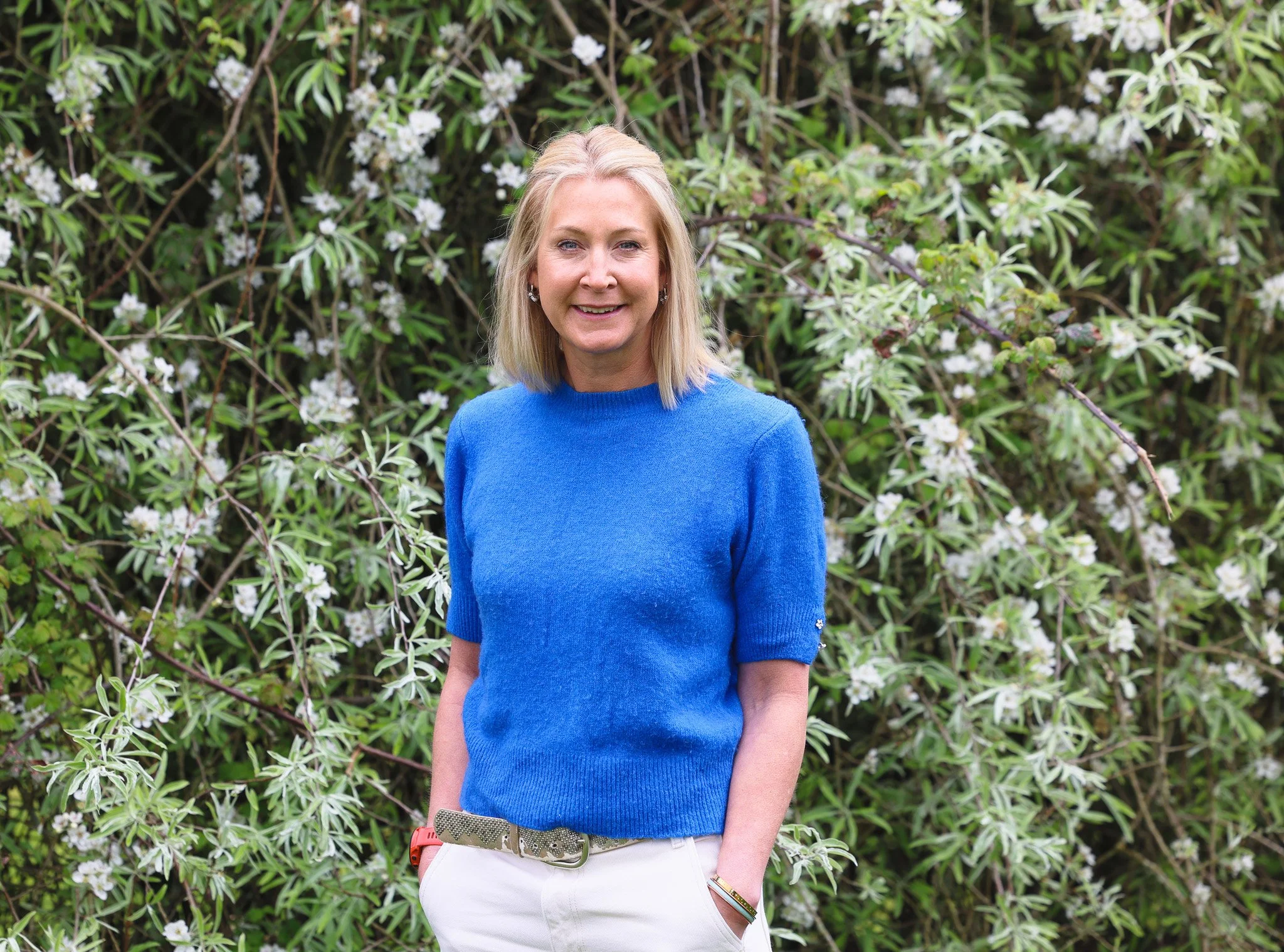 A woman with blonde hair wearing a bright blue sweater and white pants stands outdoors in front of a lush green bush with white flowers.