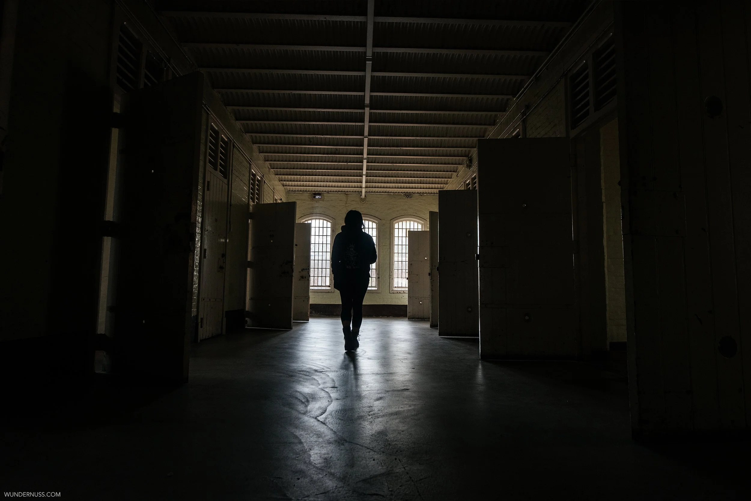 Silhouette of a person walking in a dimly lit corridor with open doors and barred windows.