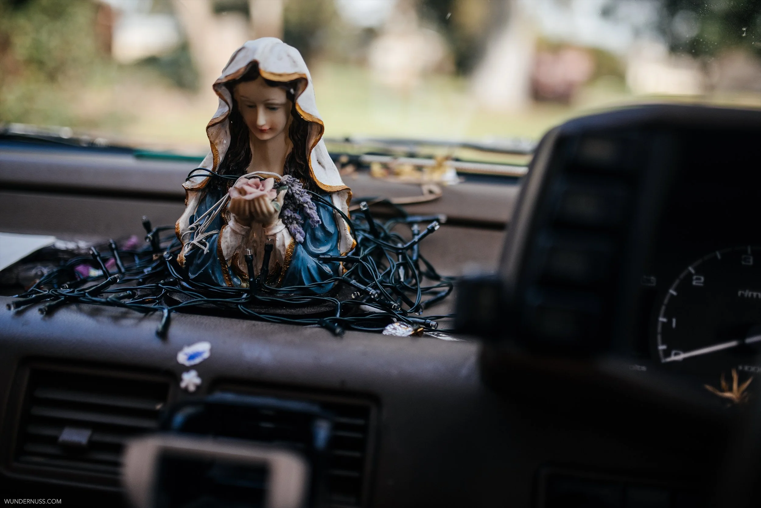 A religious figurine of a woman holding flowers is placed on a car dashboard surrounded by string lights. The dashboard and part of the speedometer are visible.