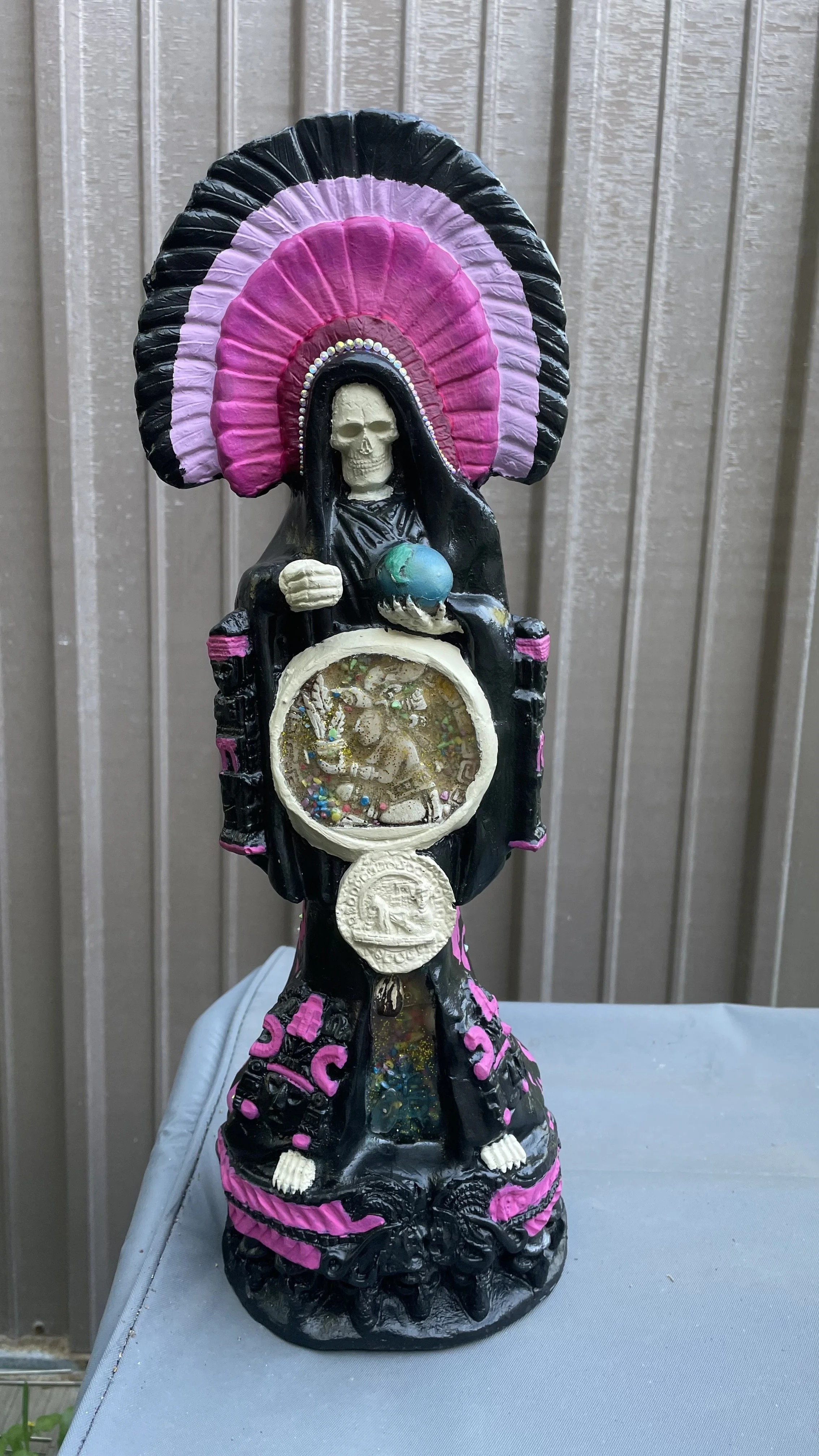 Decorative figurine of Santa Muerte with pink and black details, holding a globe and a circular inset with a scene, set against a metal panel background.