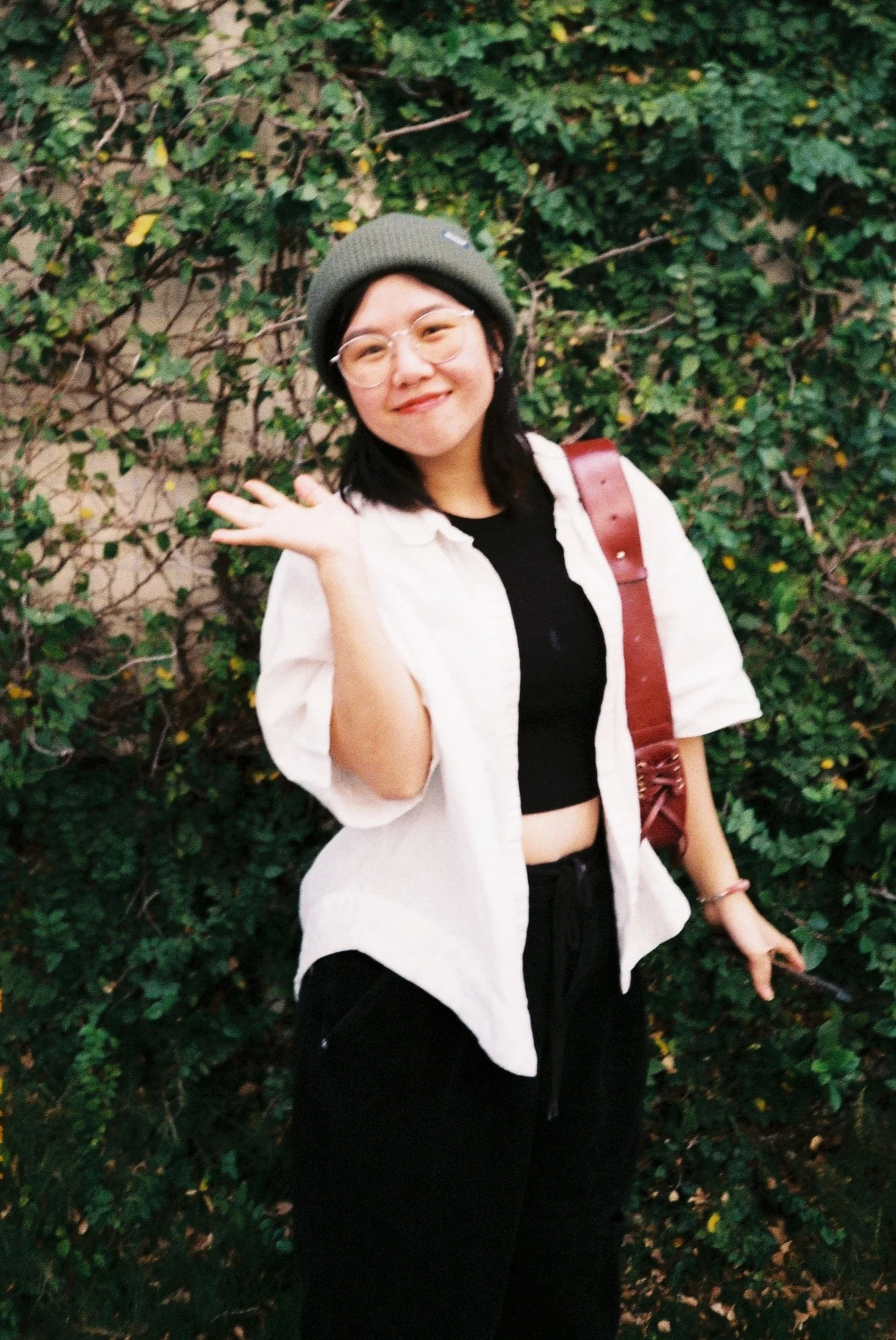 Person wearing glasses and a beanie standing in front of a greenery-covered wall, smiling and waving with one hand.