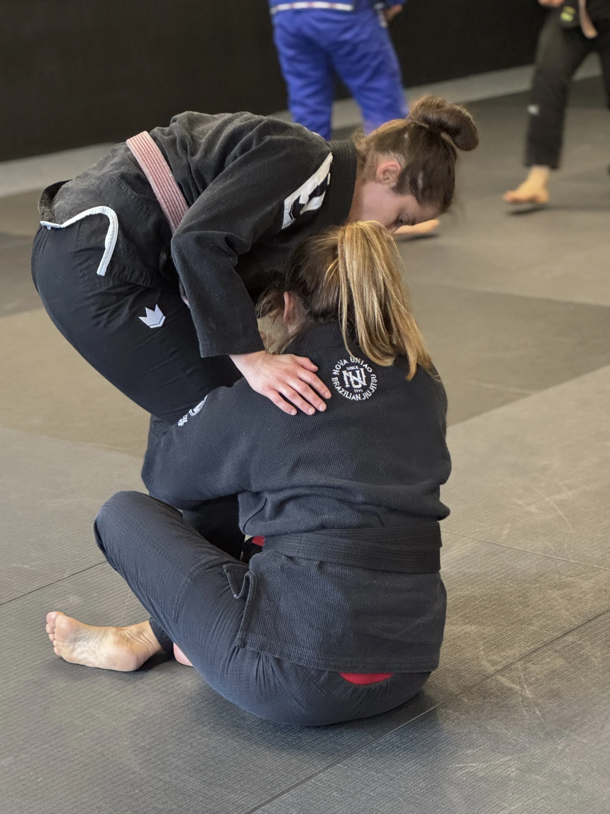 Two women practicing Brazilian Jiu-Jitsu on a mat. One woman is kneeling and leaning over the other who is seated with her legs crossed. Both are wearing black gi uniforms.
