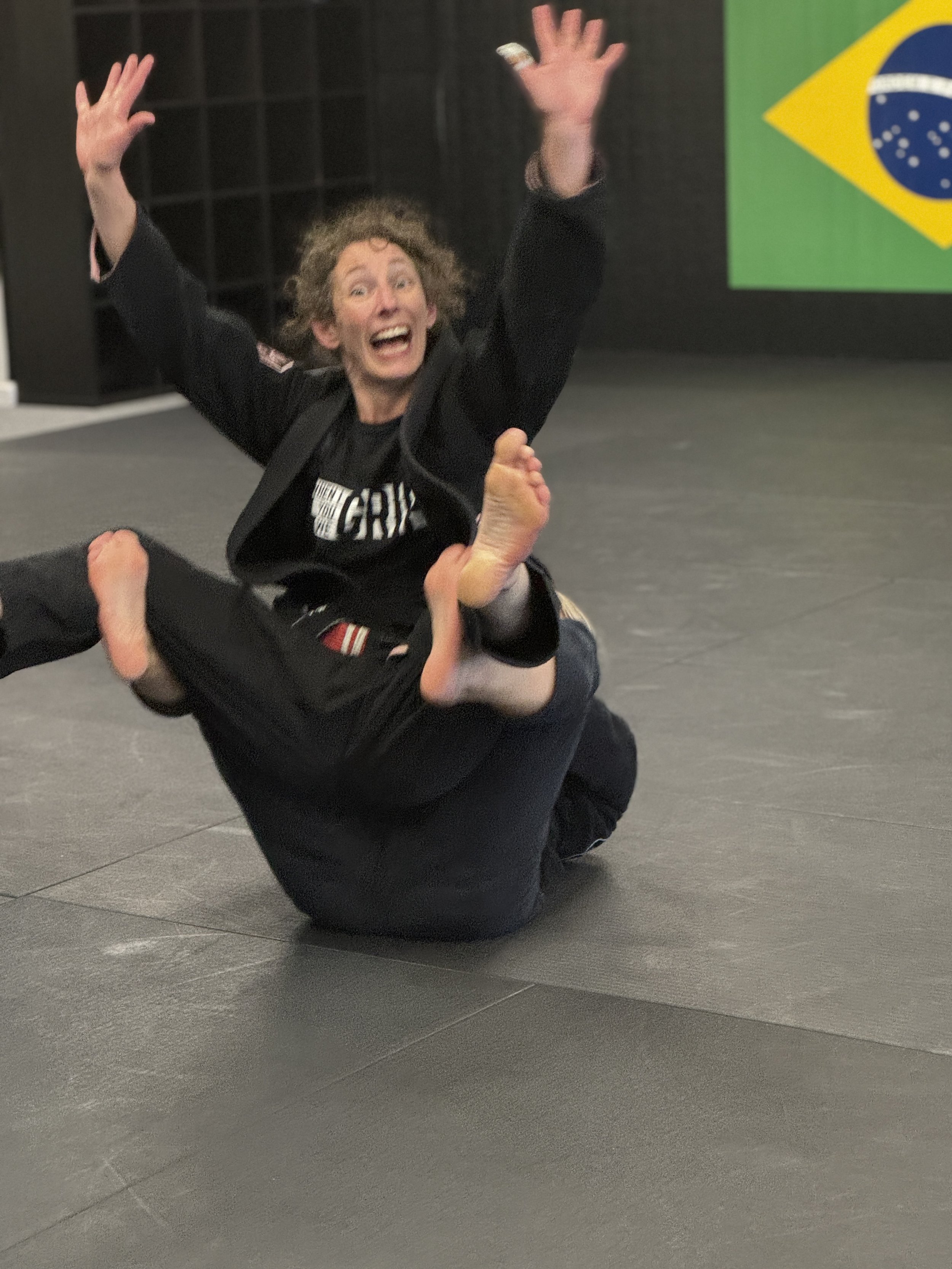 Woman in a martial arts uniform celebrates with a big smile as she holds a child in a Brazilian Jiu-Jitsu gi on the ground. Brazil flag in background.