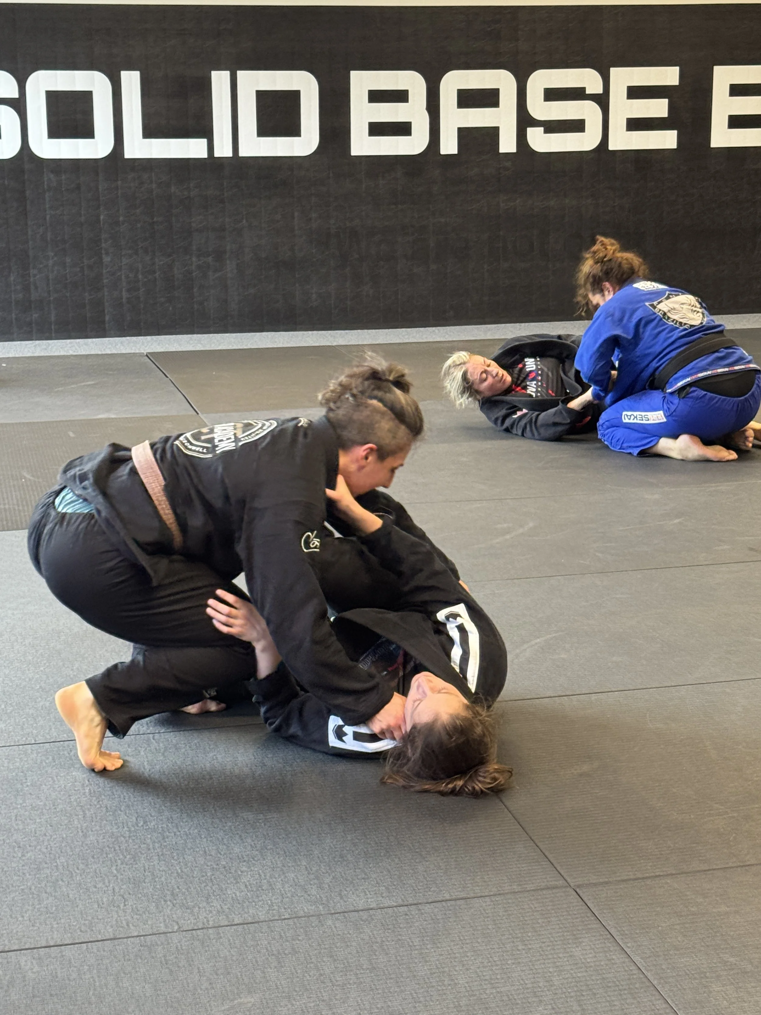 Brazilian Jiu-Jitsu practitioners training on a black mat with a black wall in the background that has large white letters spelling 'SOLID BASE'.