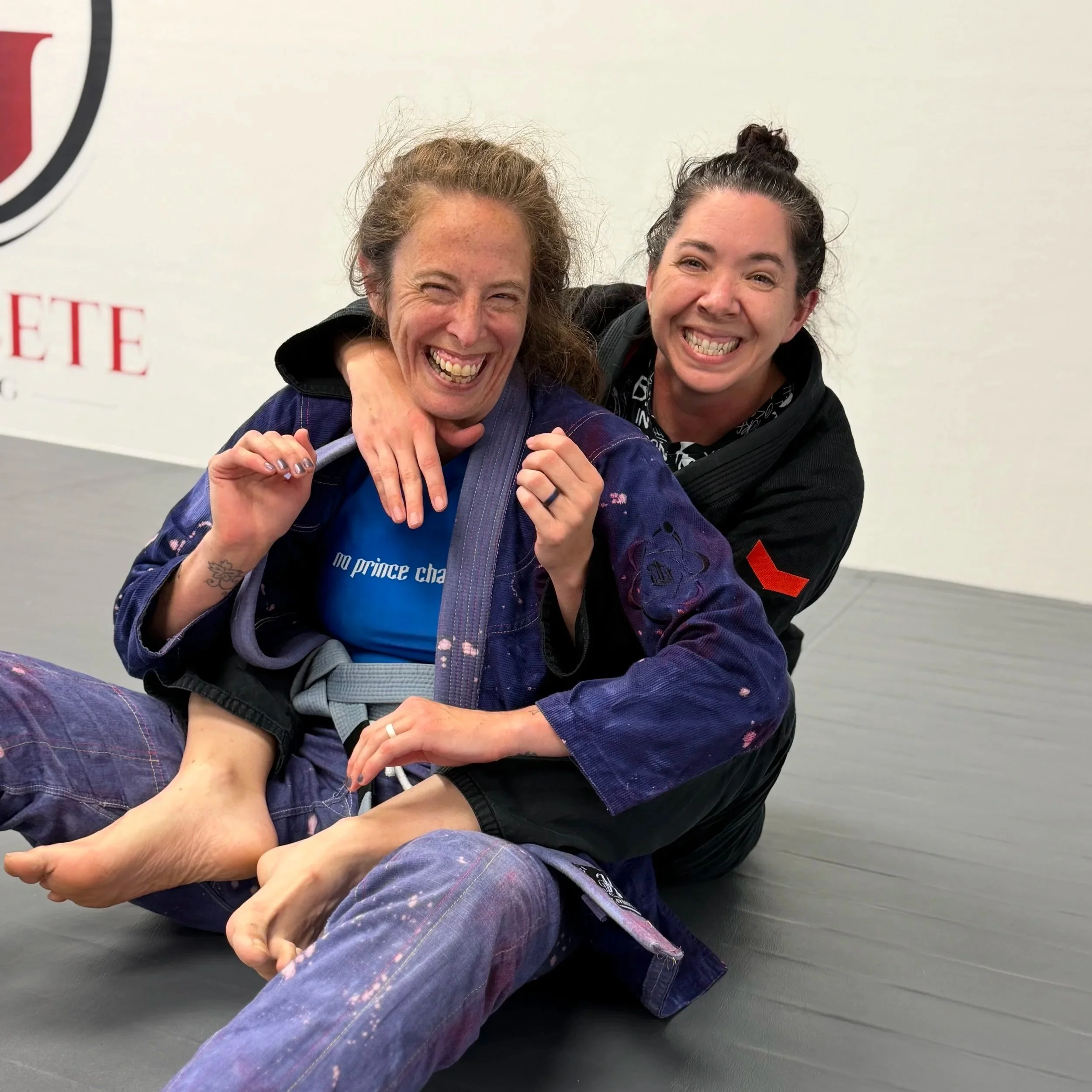Brazilian Jiu-Jitsu students practicing grappling techniques in a training facility. A woman is on her back with a man on top, engaging in a ground fighting position, with several other students training in the background.