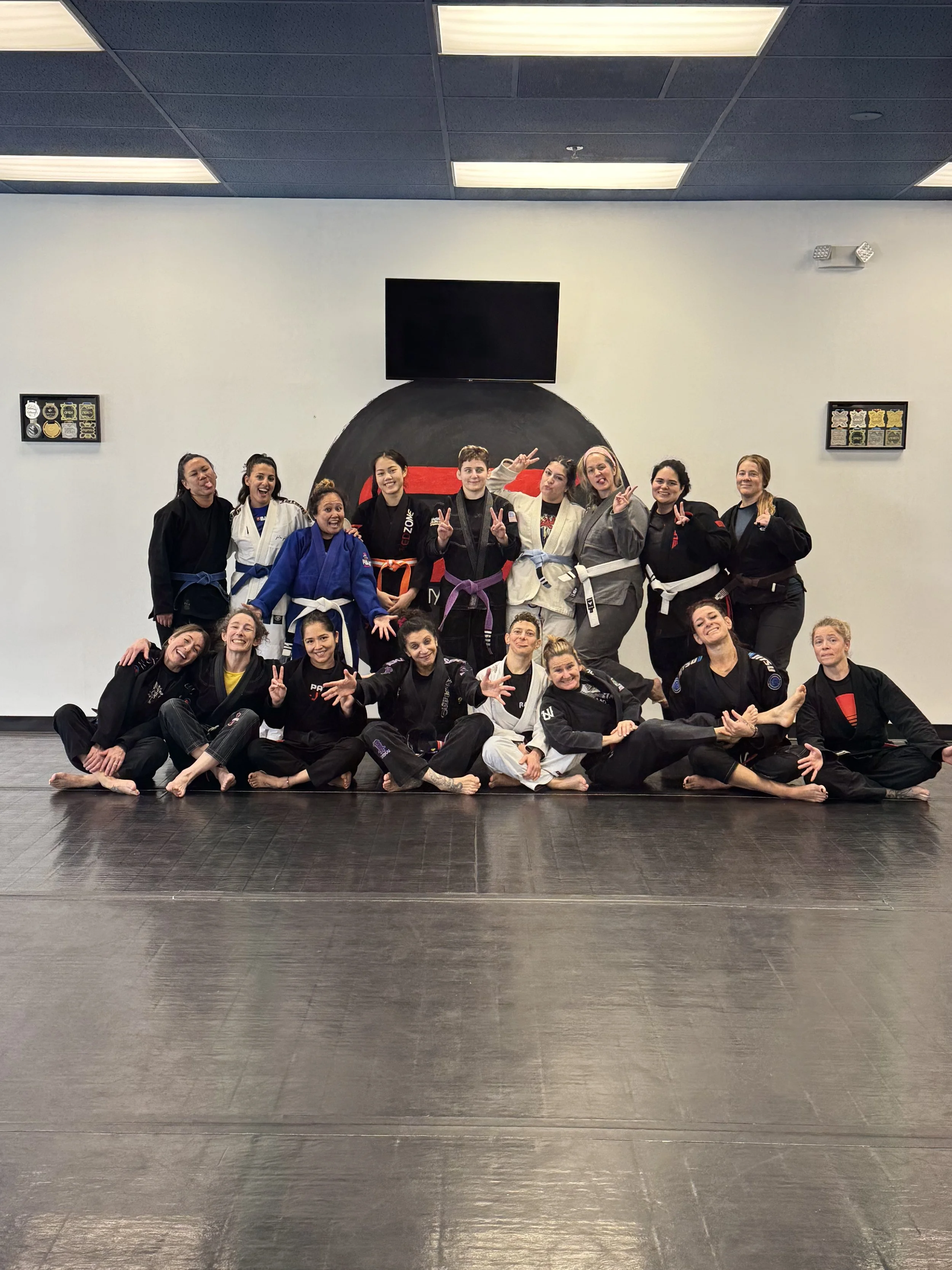 Two women practicing Brazilian Jiu-Jitsu on a black mat, with one woman on the ground holding a wrist lock, and the other woman leaning over her, applying pressure. Two other women in the background are practicing grappling.