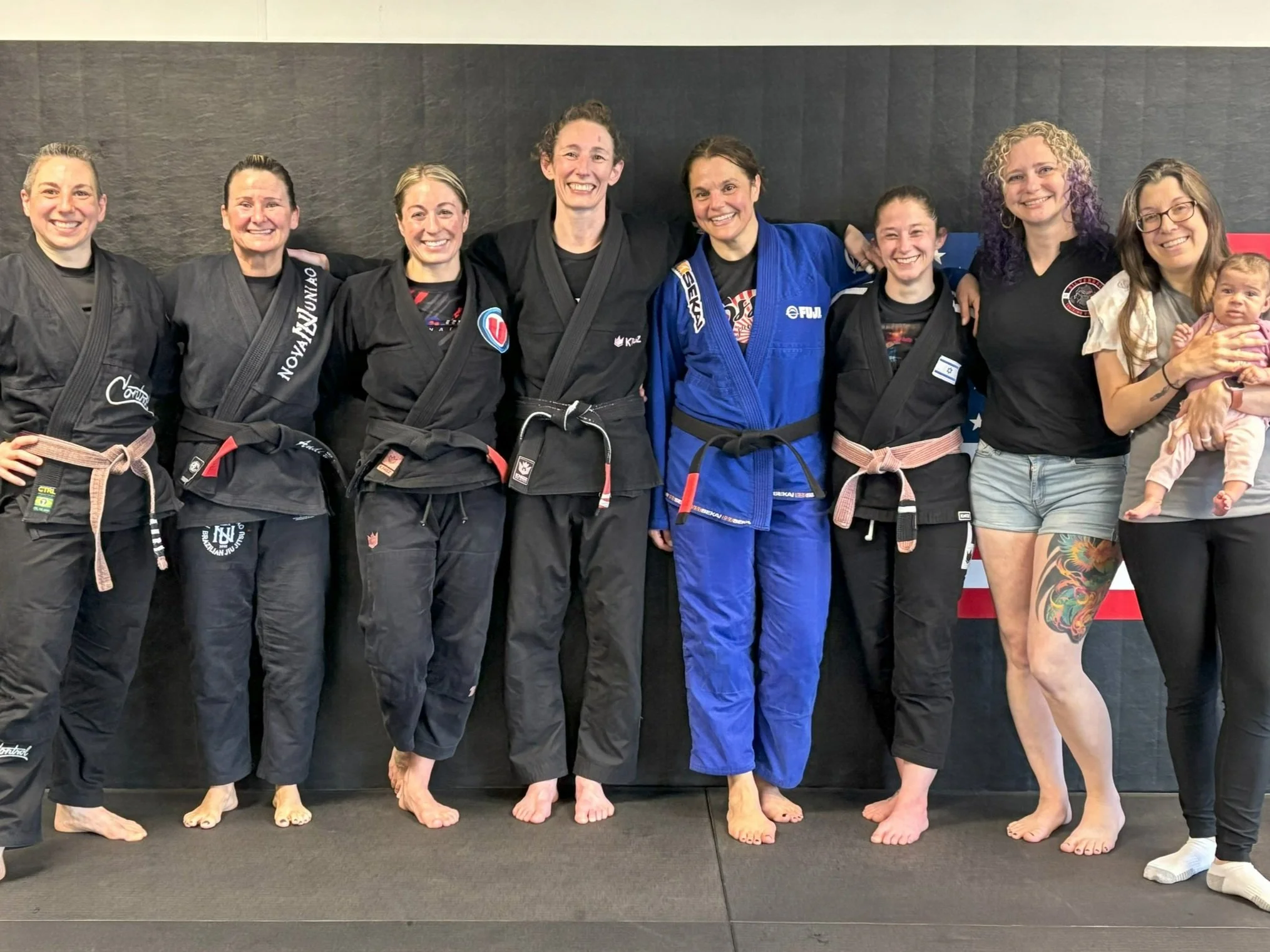 Group of women in Brazilian Jiu-Jitsu gis standing together in a gym, smiling, with one woman holding a baby.