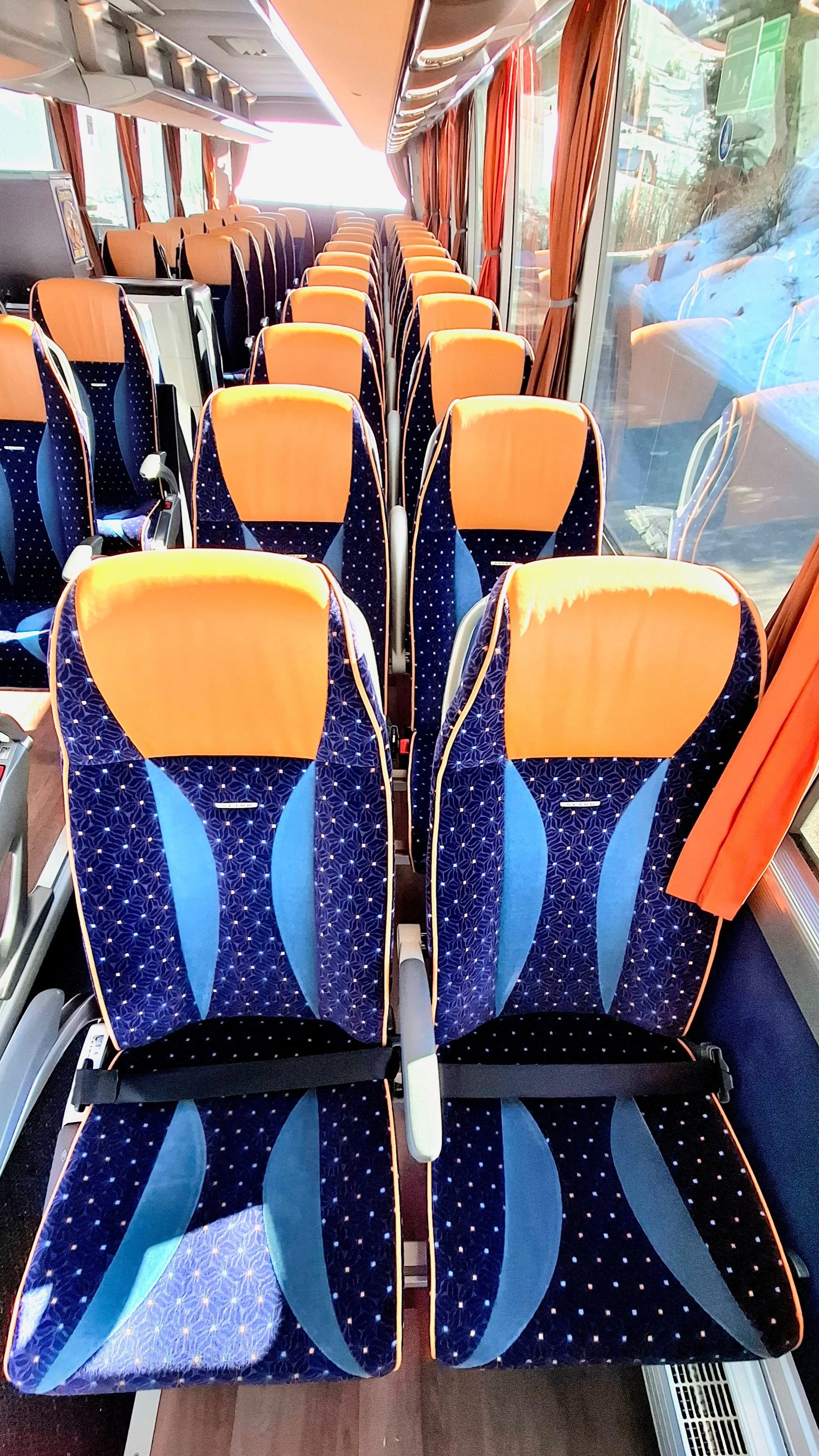 Innenansicht eines Bus mit blaue und orange Sitze, Fenster mit roten Vorhängen, Blick nach vorne.