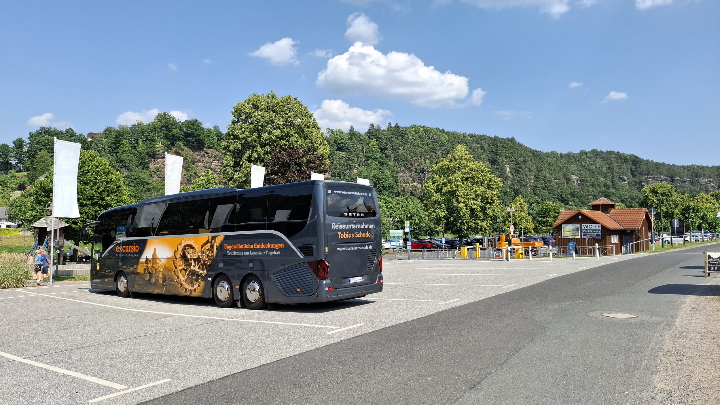 Ein schwarzer Bus mit Werbung für Tourismus steht auf einem Parkplatz mit Bäumen und Hügel im Hintergrund, bei sonnigem Wetter.