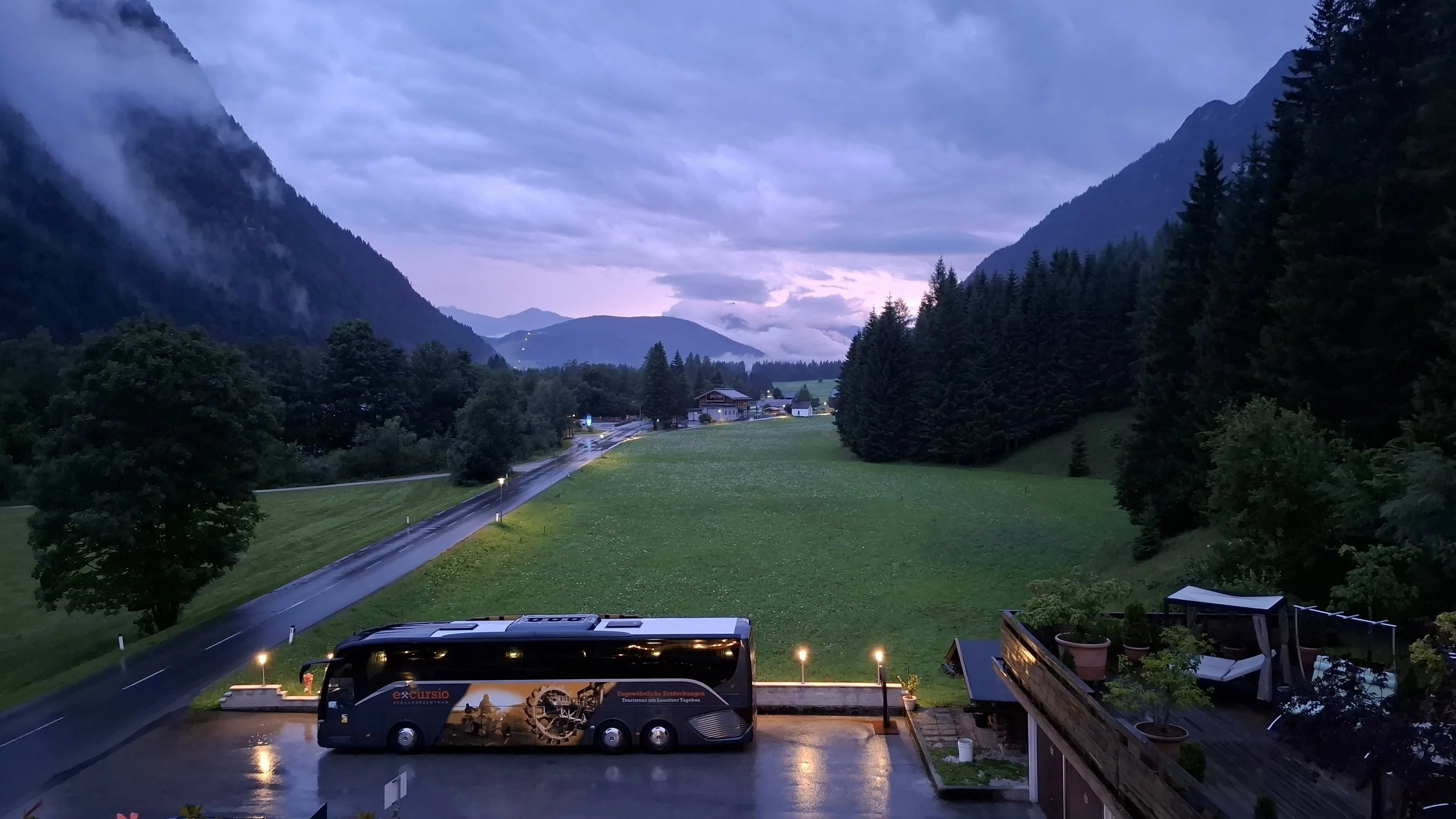 Blick auf eine nasse Straße, grüne Wiesen, eine große Bus mit der Aufschrift 'excursio', und einen Balkon mit Pflanzen bei Dämmerung in einer bergigen Landschaft.