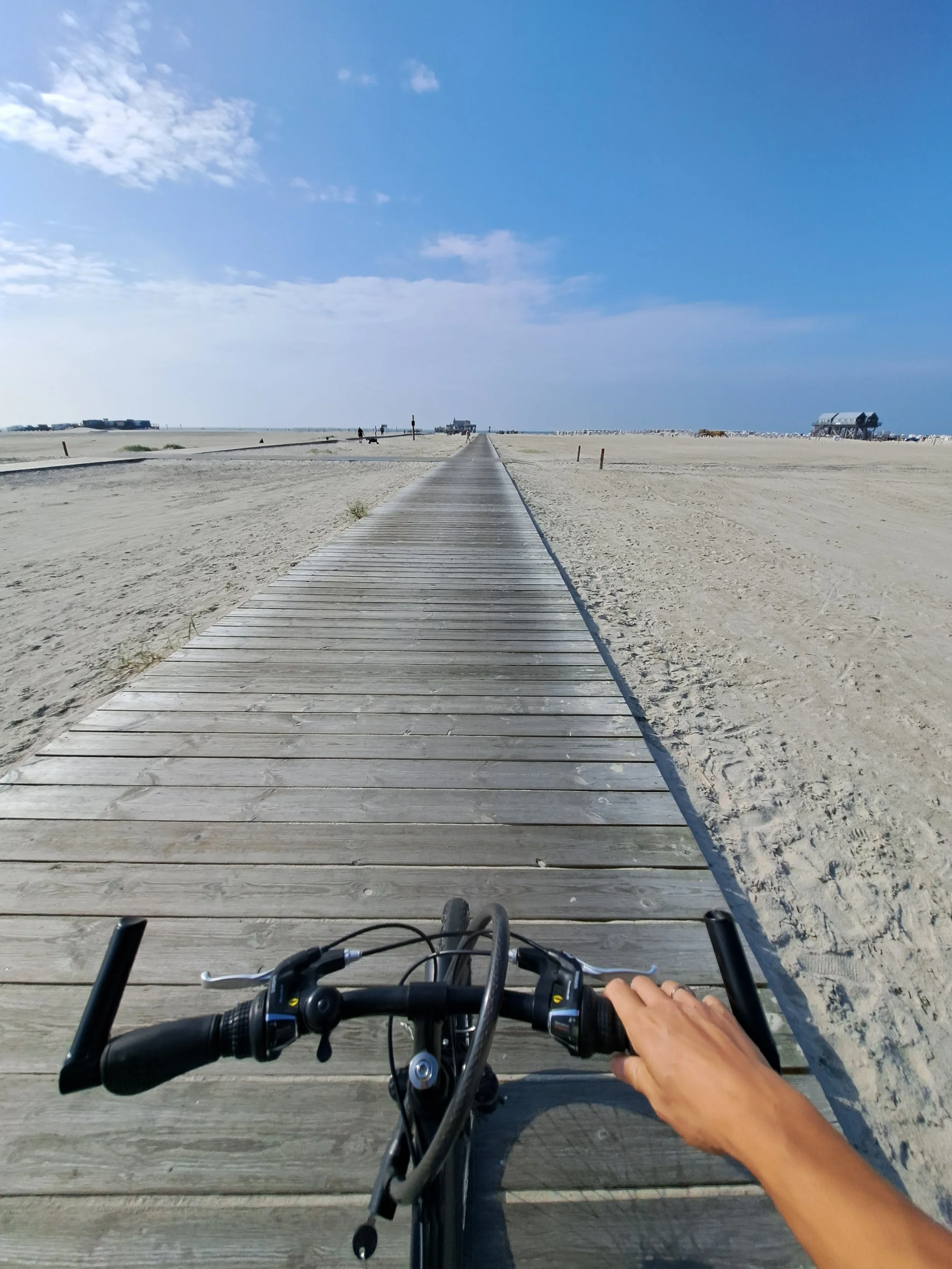 Blick vom Fahrrad auf einen Holzsteg am Strand unter blauem Himmel.