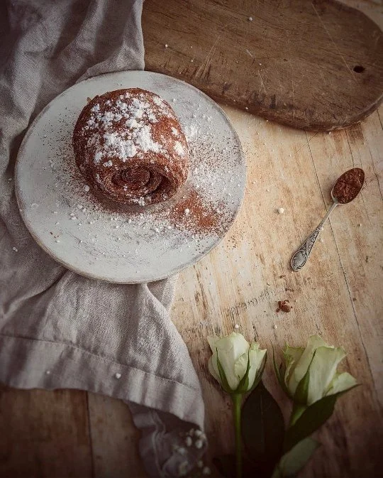A cinnamon roll dusted with powdered sugar on a white plate, placed on a beige cloth on a wooden table, with a spoon of cinnamon, two white roses, and a wooden cutting board nearby.