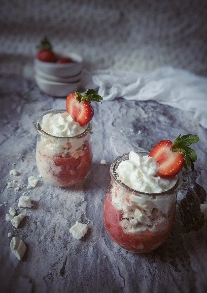 Two glass jars filled with strawberry dessert topped with whipped cream and fresh strawberry slices with mint leaves, placed on a textured gray surface with scattered crumbs and a white napkin in the background.