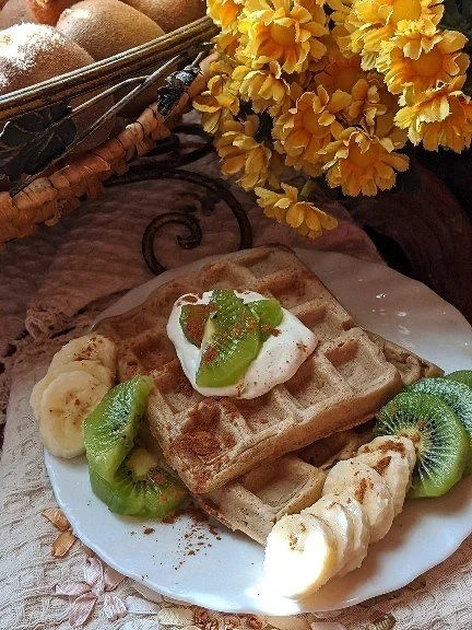 A plate with waffles topped with whipped cream, sliced banana, kiwi, and a drizzle of honey, with additional banana and kiwi slices on the side. There are yellow flowers and a basket with apples and other fruits in the background.