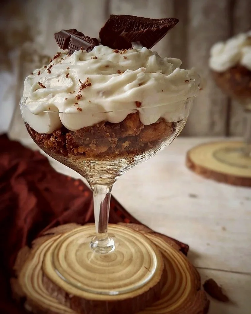 A dessert in a glass with layers of crumbled cookies, topped with whipped cream, chocolate shavings, and a piece of chocolate cake.