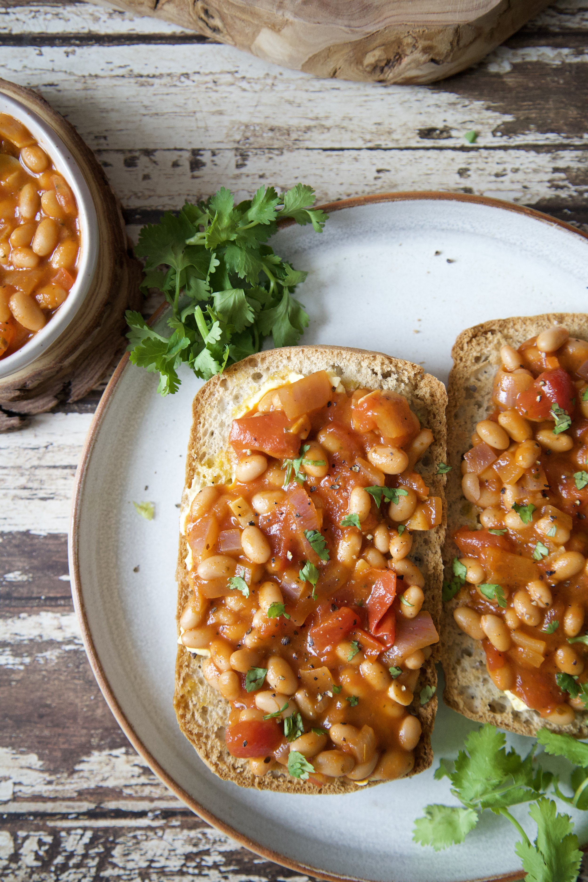 Two pieces of toasted bread topped with baked beans, diced tomatoes, and herbs on a white plate, with fresh cilantro garnish and a bowl of baked beans in the background on a rustic wooden table.