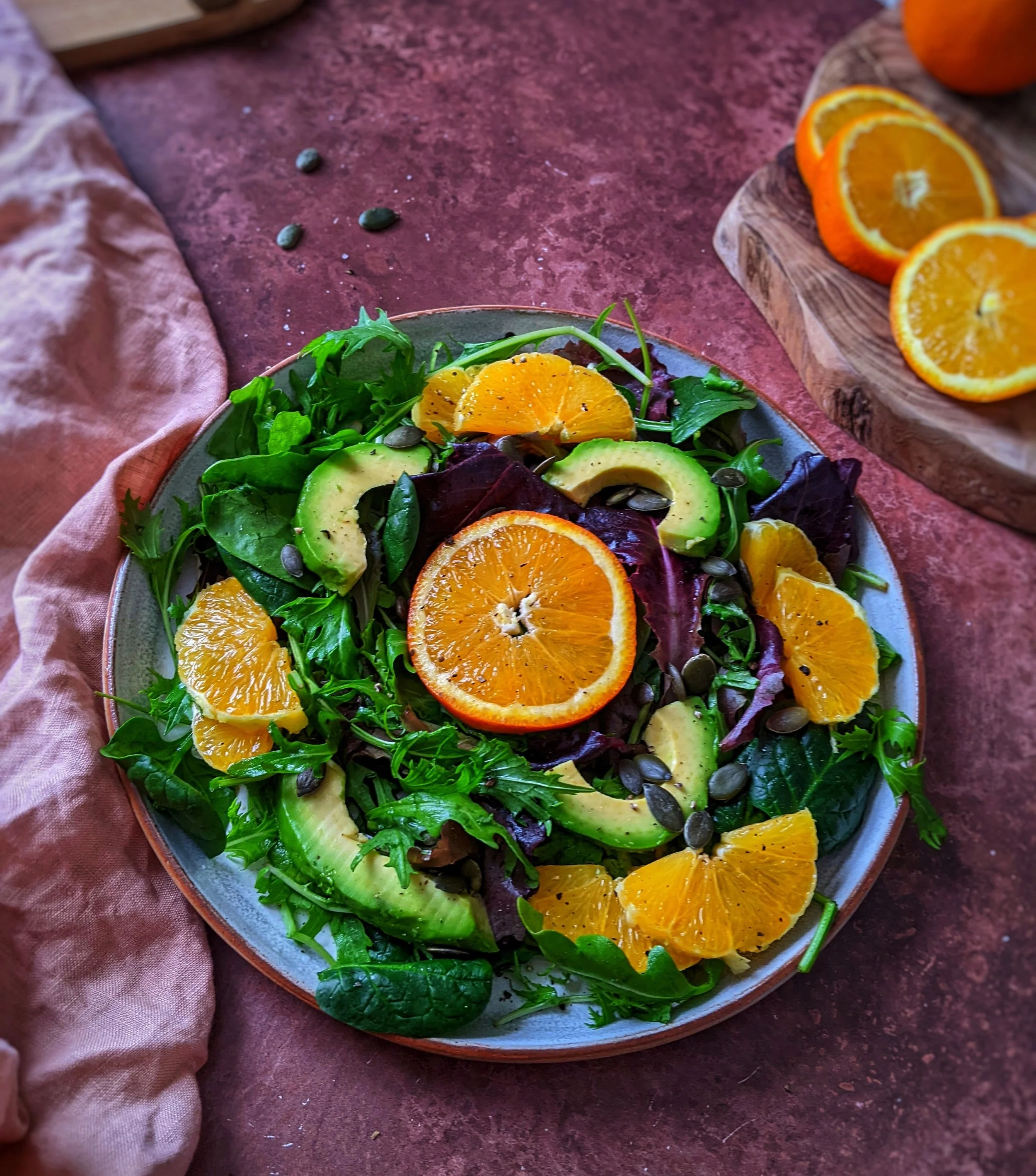 A colorful salad with mixed greens, sliced avocados, orange slices, and pumpkin seeds on a gray plate, with additional orange slices on a wooden cutting board nearby.