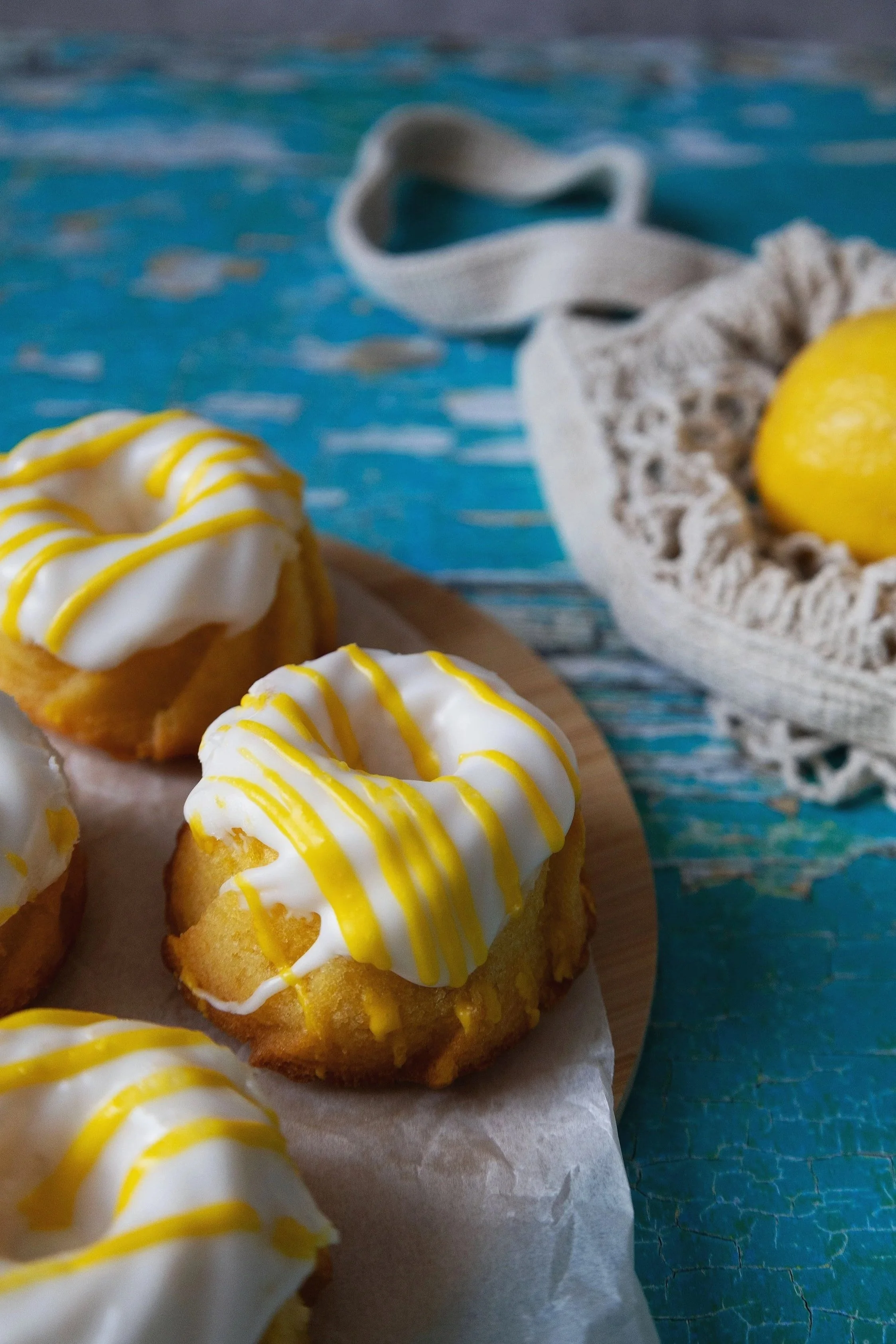 Close-up of lemon cupcakes with white icing and yellow drizzle on a wooden surface, with a lemon and a cloth bag in the background.
