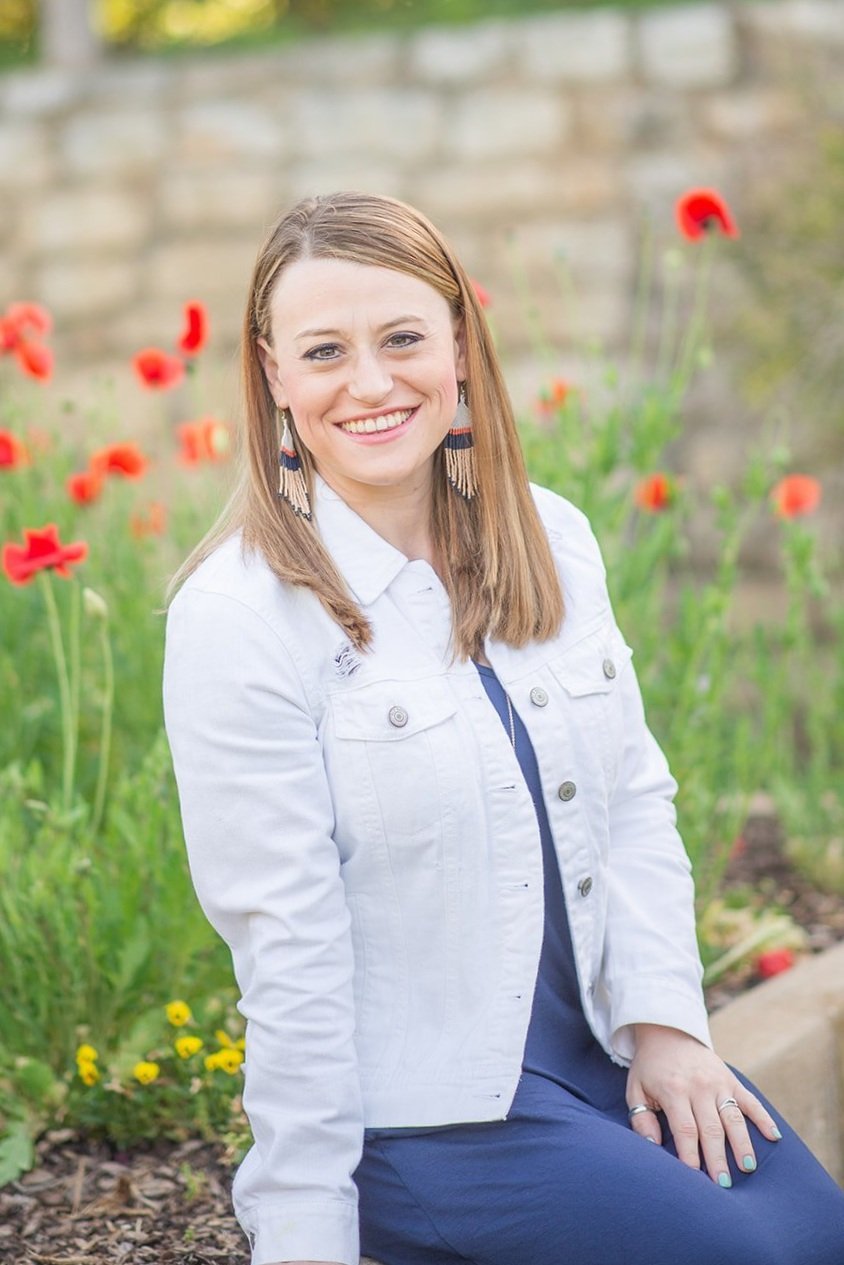 Ashley Hardy LPC Headshot, Ashley is smiling. and sitting amongst pretty red flowers