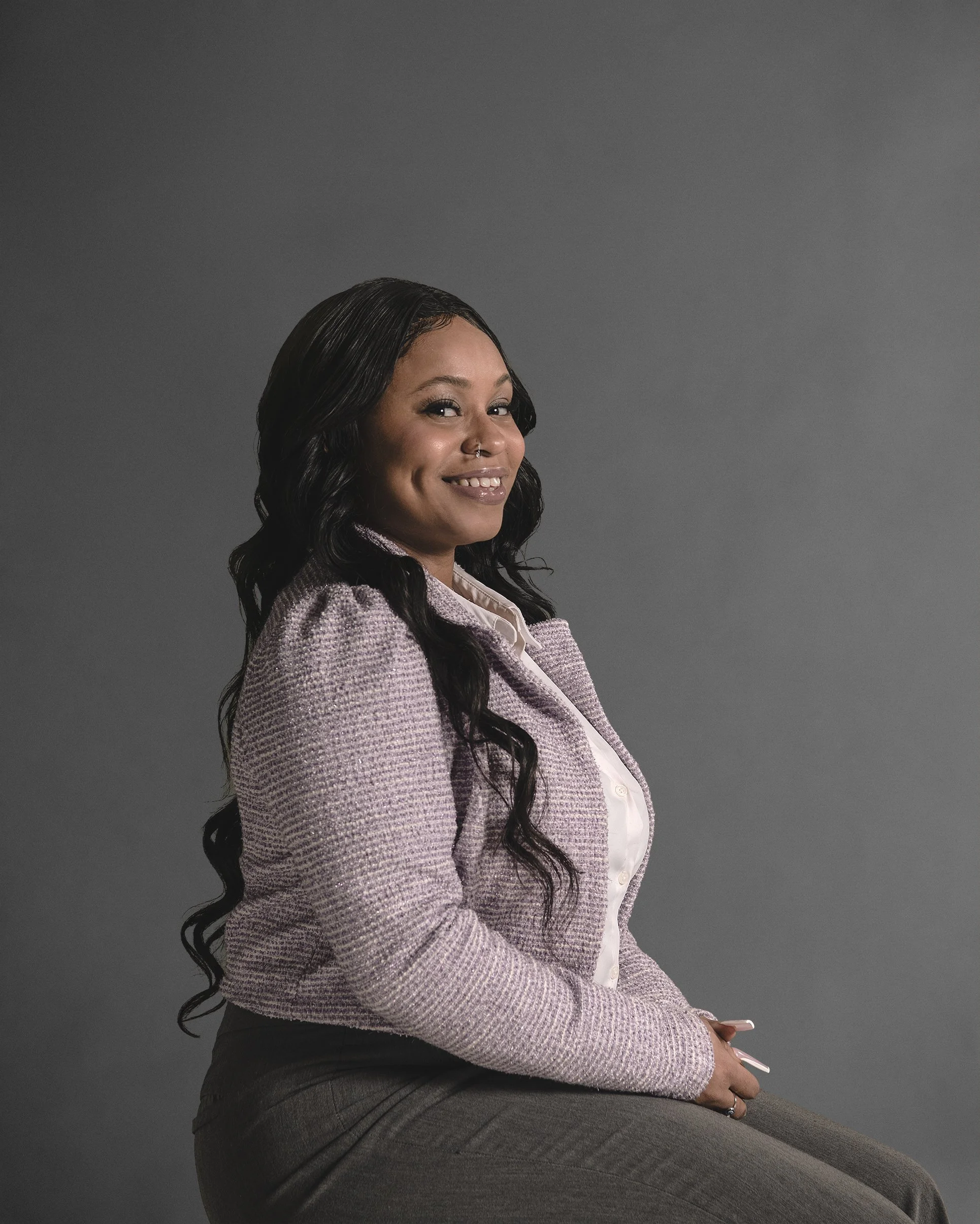 A woman with long black wavy hair and a nose ring smiling, seated against a plain gray background, wearing a light purple textured blazer and a white blouse.