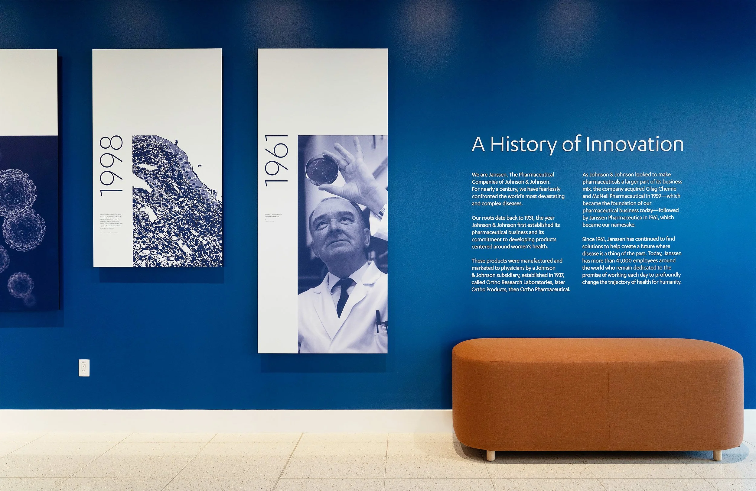 A blue wall with an informational display titled 'A History of Innovation.' The display includes historical images and text about Johnson & Johnson's pharmaceutical legacy from 1931 onward. To the right, there is an orange, curved bench.