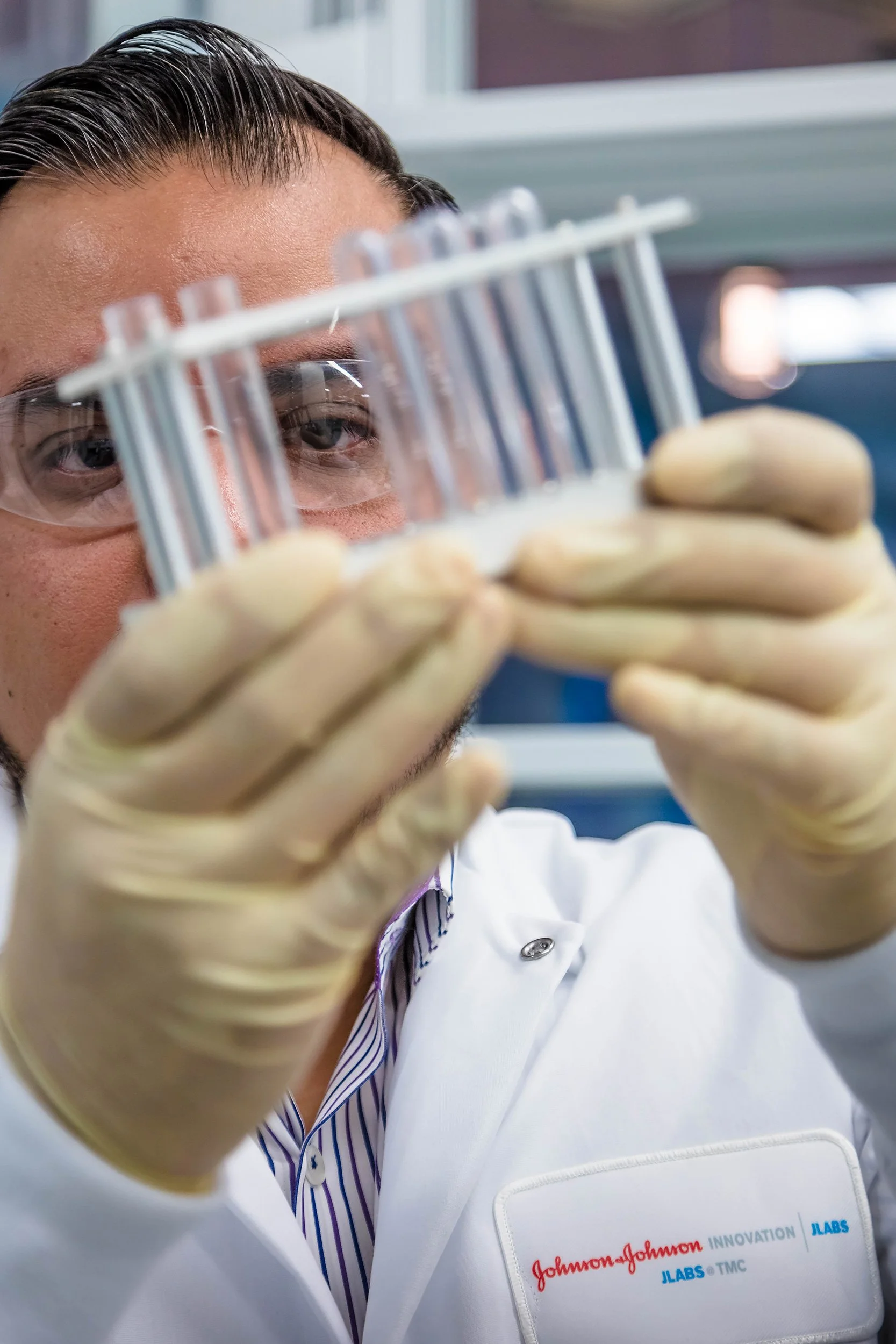 Close-up of a scientist working in a lab, wearing safety glasses and yellow gloves, holding a test tube with a focus on their face and lab coat with Johnson & Johnson branding.