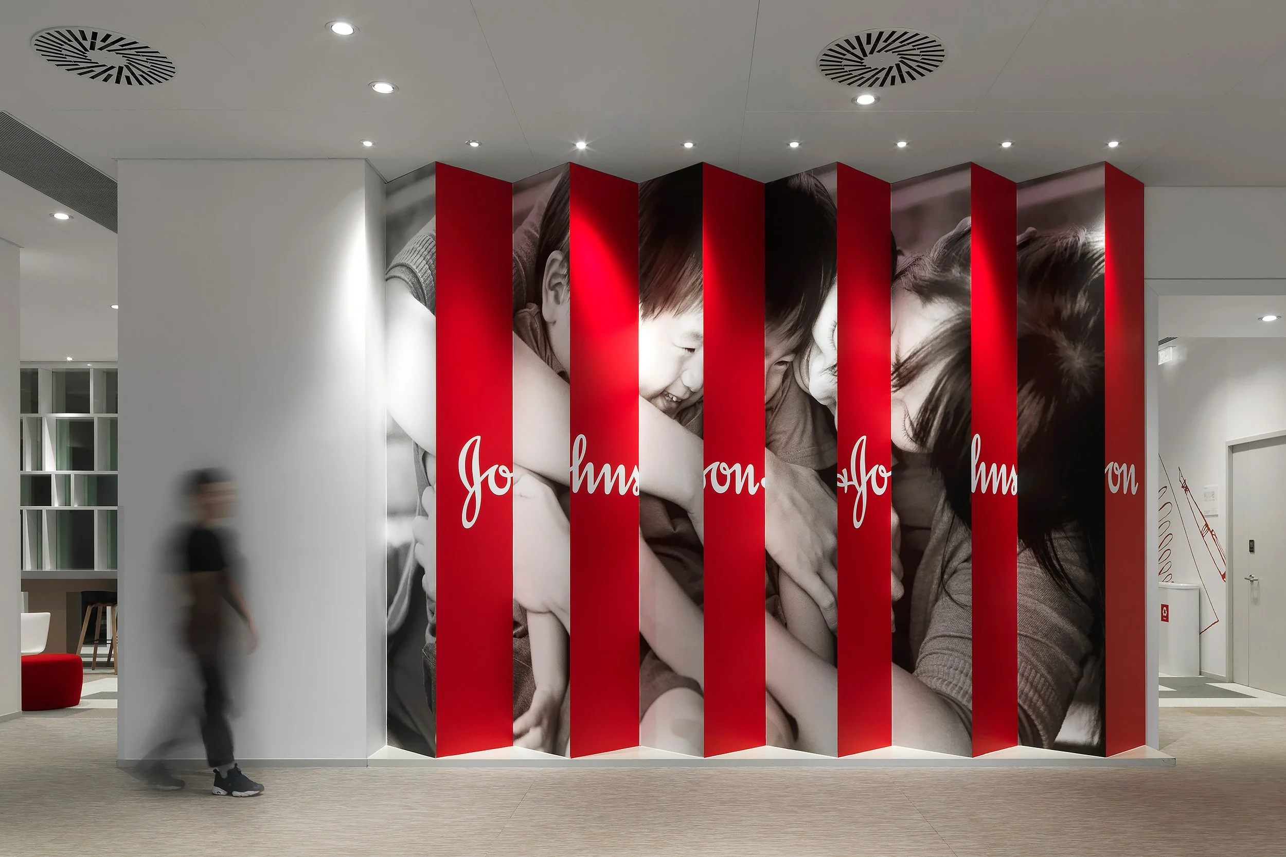 Interior view of a modern building with a large black and white photographic mural of children, segmented by red vertical panels with white script. A person is blurred walking past, and there are ceiling lights and ventilation fixtures.