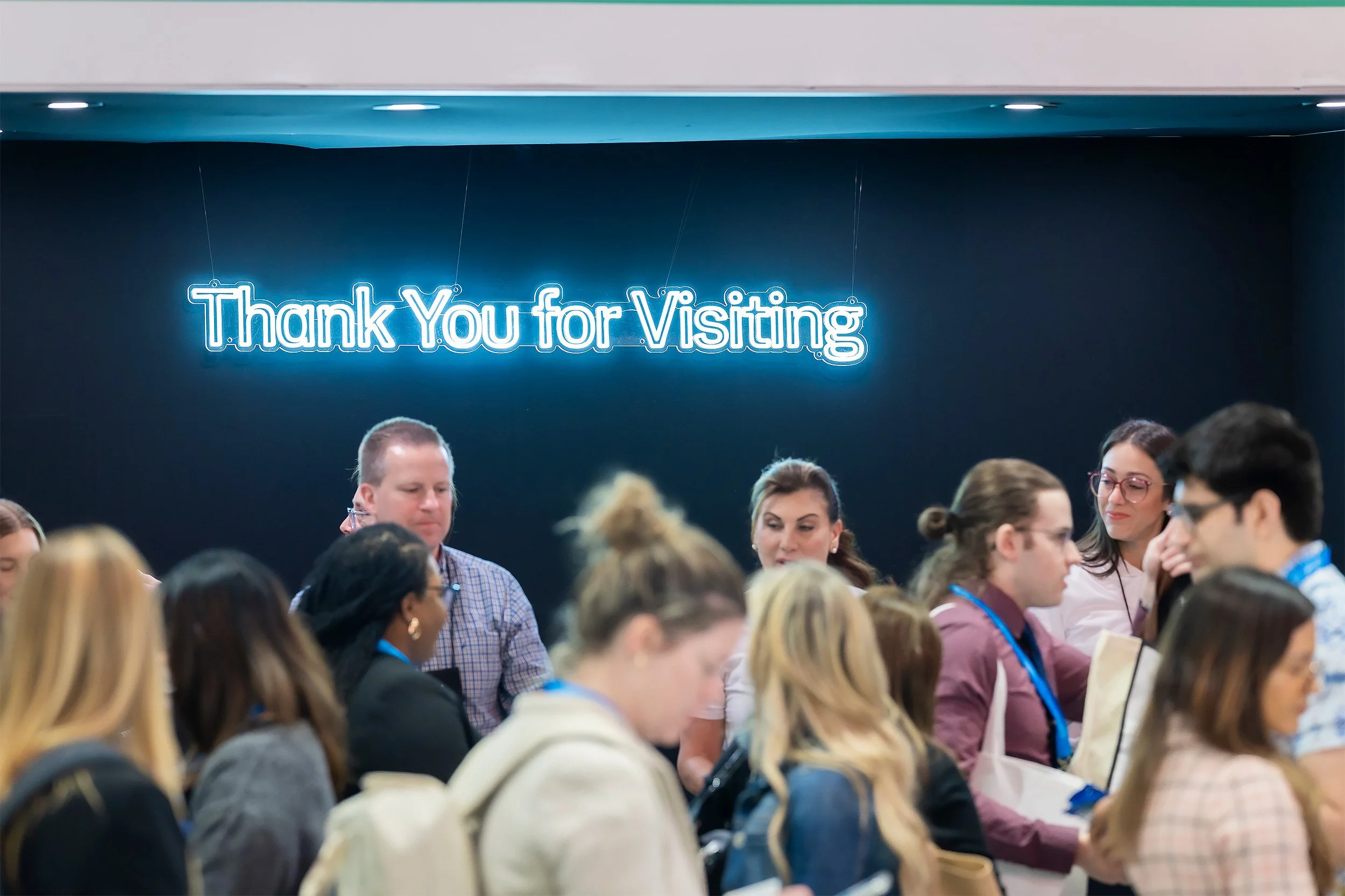 A group of people seated closely together at a conference or seminar, with a blue neon sign above them that says 'Thank You for Visiting'.